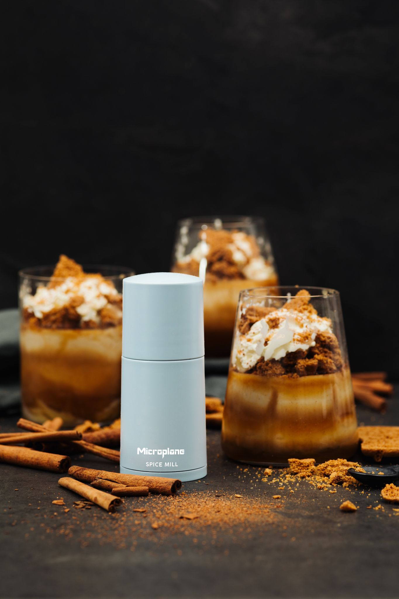 Desserts in Gläsern mit Schlagsahne und Krümeln, neben einer Gewürzmühle und Zimtstangen auf dunkler Oberfläche.