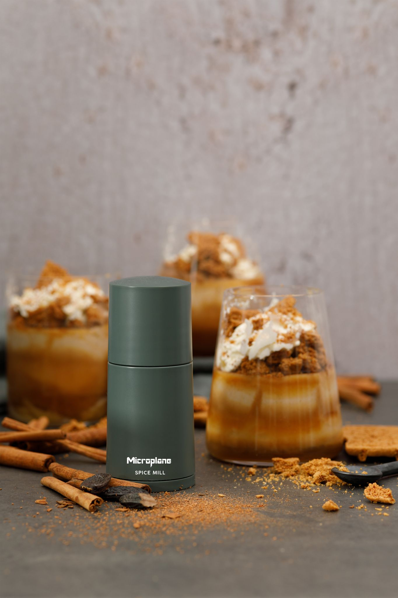 Eine grüne Gewürzmühle, drei Desserts in Gläsern mit Sahne und Krümeln, Zimtstangen und Gewürzen auf dunkler Oberfläche.