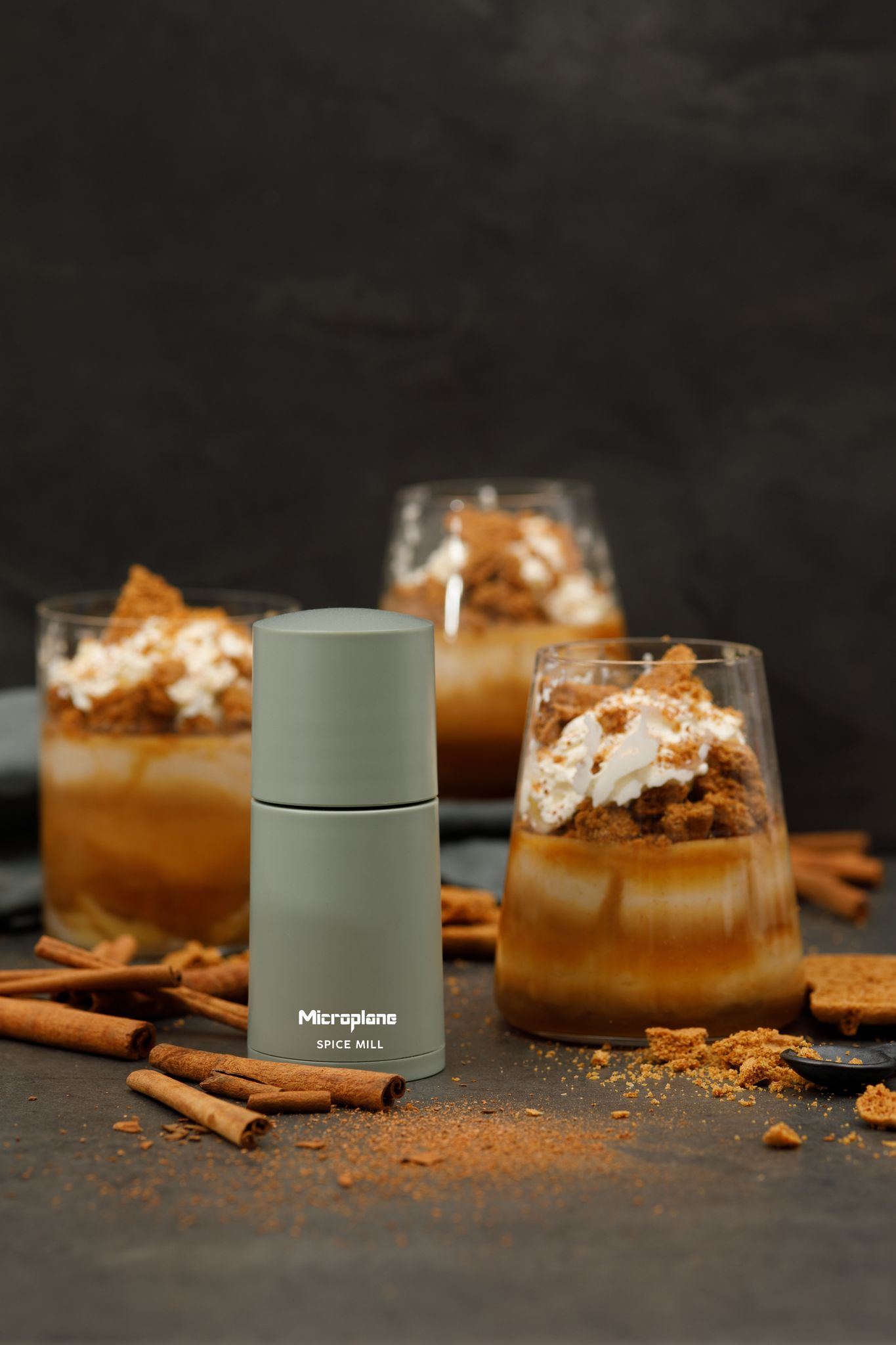Gewürzmühle, Desserts in Gläsern, Zimtstangen und Krümel auf dunkler Oberfläche. Die Desserts haben Schlagsahne und Kekse.