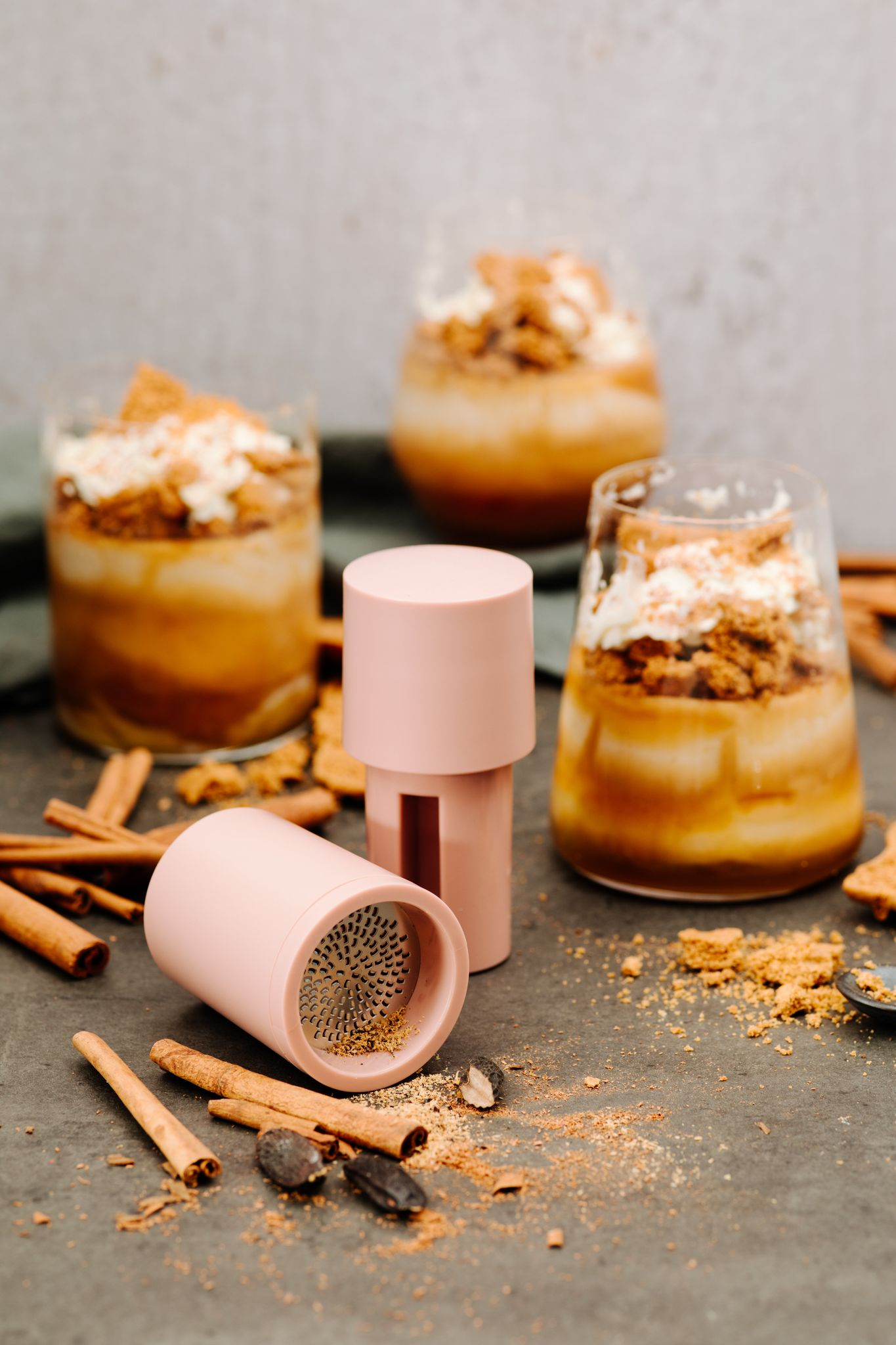 Drei Desserts in Gläsern, rosa Gewürzmühlen, Zimtstangen und Keksbrösel auf einer grauen Oberfläche.