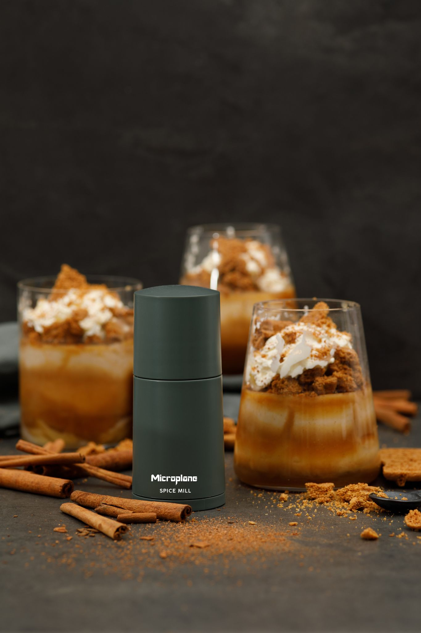 Eine Gewürzmühle steht vor Desserts in Gläsern, mit Zimtstangen und Krümeln drum herum, auf dunkler Oberfläche.