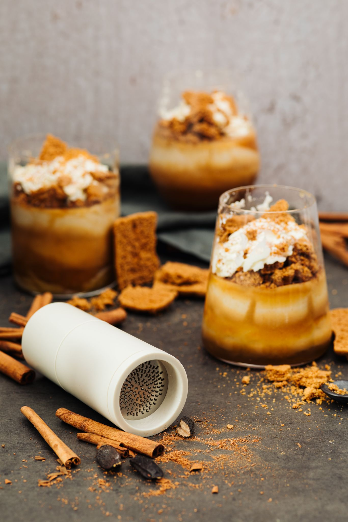 Desserts in Gläsern mit Sahne und Keksen. Zimtstangen, eine Reibe und Krümel sind verstreut.