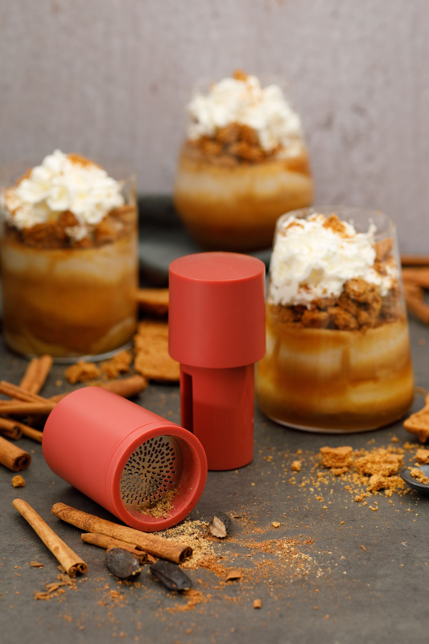 Dessertgläser mit Schlagsahne, Keksen und Zimtstangen. Roter Grinder im Vordergrund, auf dunkler strukturierter Oberfläche.