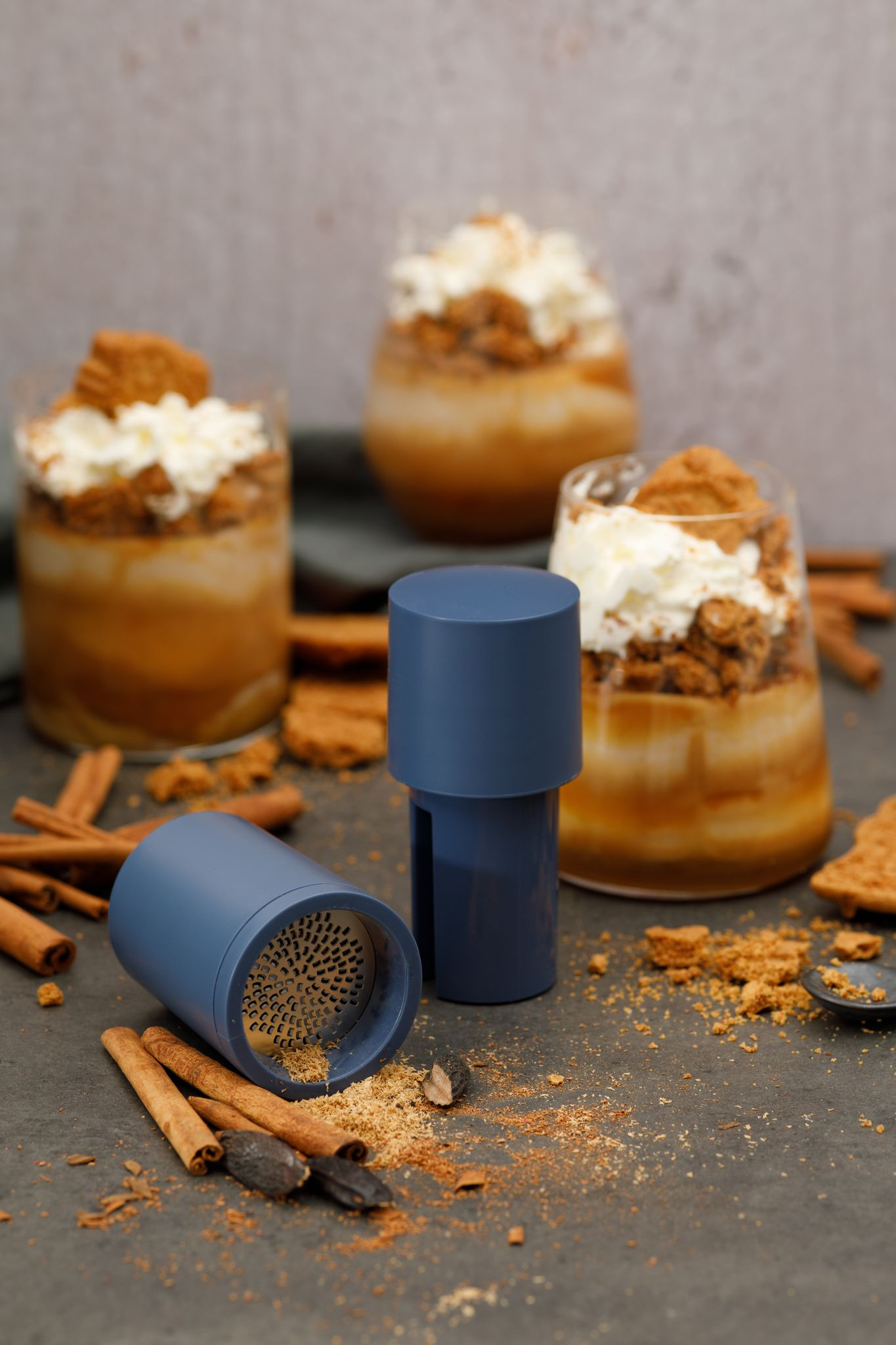 Desserts in Gläsern mit Schlagsahne, Keksen und Gewürzen. Blauer Grinder und Zimtstangen liegen auf grauer Oberfläche.