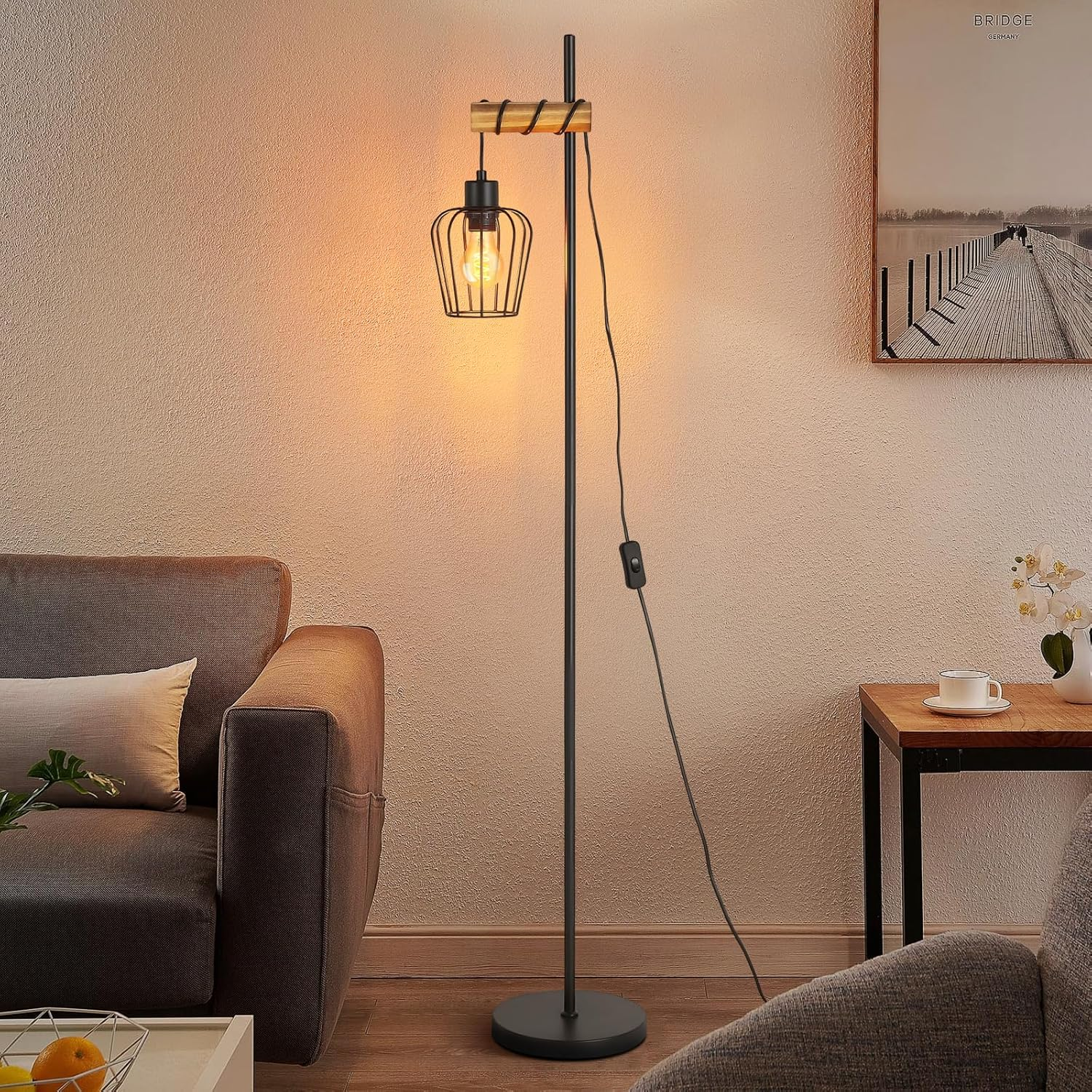 Una lampada da terra con paralume a gabbia, un divano, un tavolino con tazza e un quadro con ponte.