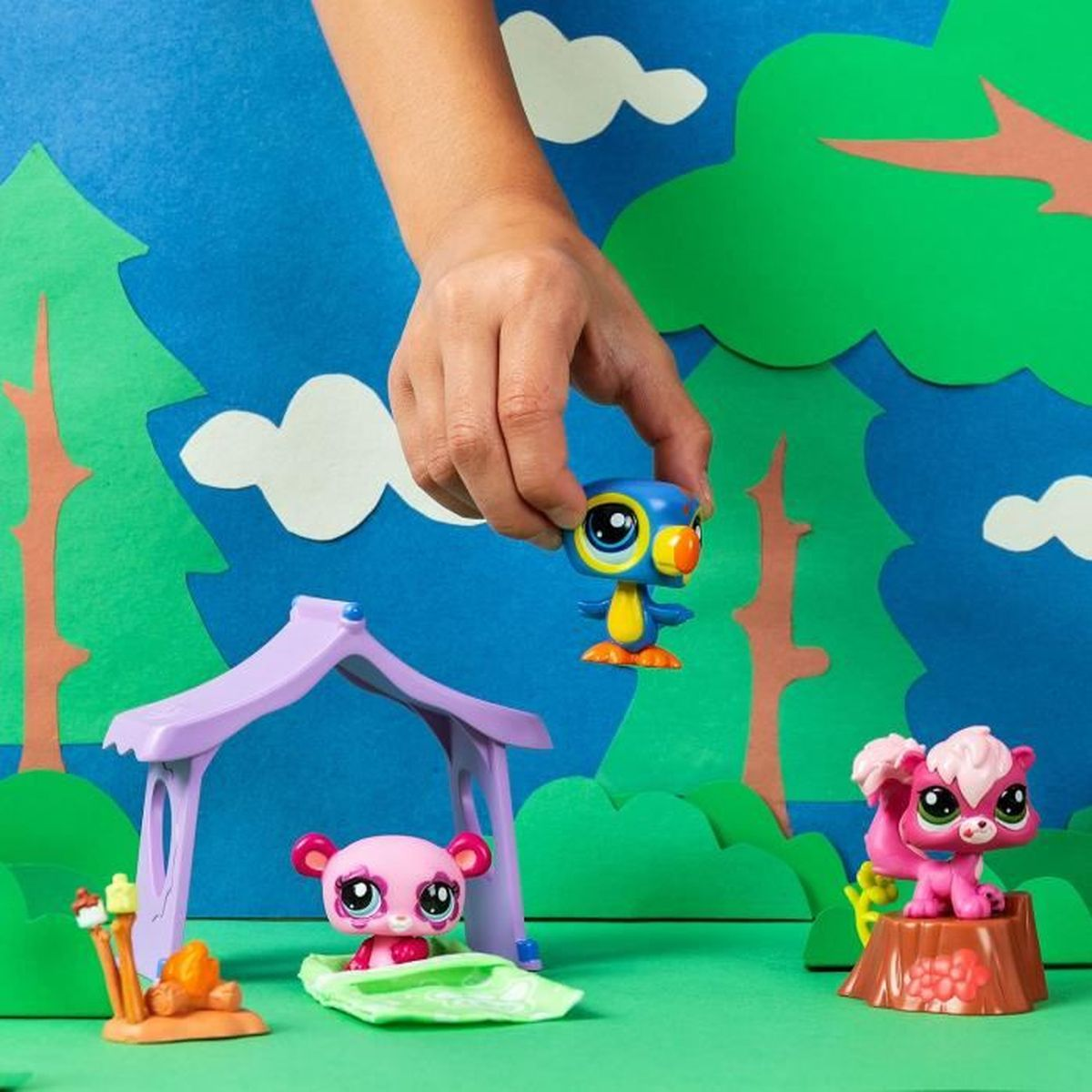 Eine Hand hält ein blaues Vogelspielzeug. Rosa Spielzeuge eines Bären und Eichhörnchens sind in einem Zelt und auf einem Baumstumpf. Hintergrund ist eine Waldszene.