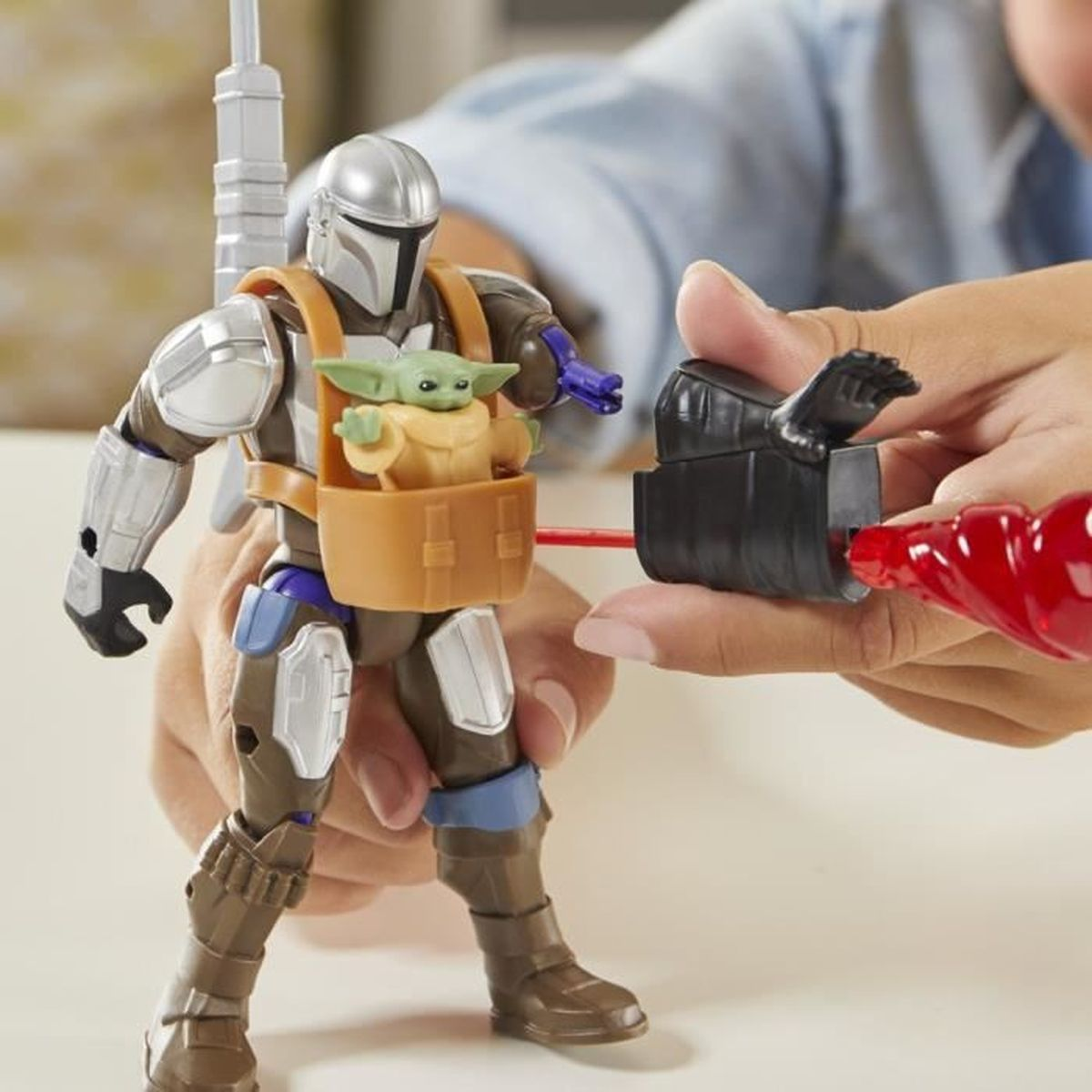 Eine Mandalorianer-Actionfigur mit Baby Yoda in einer Trage wird von einer Person gehalten. Ein roter Strahl wird auf die Figur geschossen.