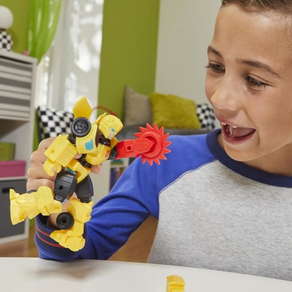 Un niño sonríe, sosteniendo un robot de juguete amarillo con una sierra roja. Un pequeño bloque amarillo está en la mesa. Fondo: habitación con muebles.