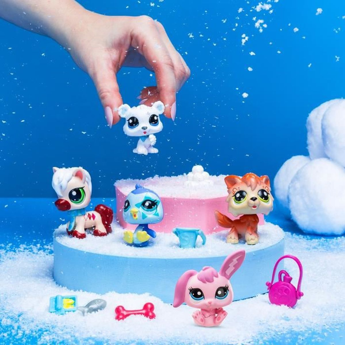 Eine Hand hält ein Eisbären-Spielzeug über anderen Tier-Spielzeugen auf einer schneebedeckten Plattform. Rosa Hase, orangefarbene Katze, Pferd und blauer Vogel sind vorhanden.