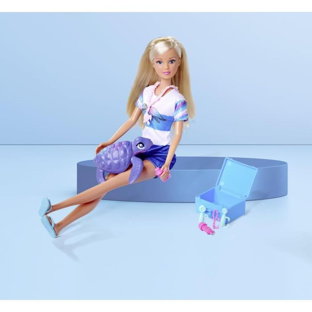 Blonde Puppe mit einer lila Schildkröte. Rechts befindet sich eine blaue Box mit Zubehör. Die Puppe trägt blaue Shorts und ein weißes Oberteil.