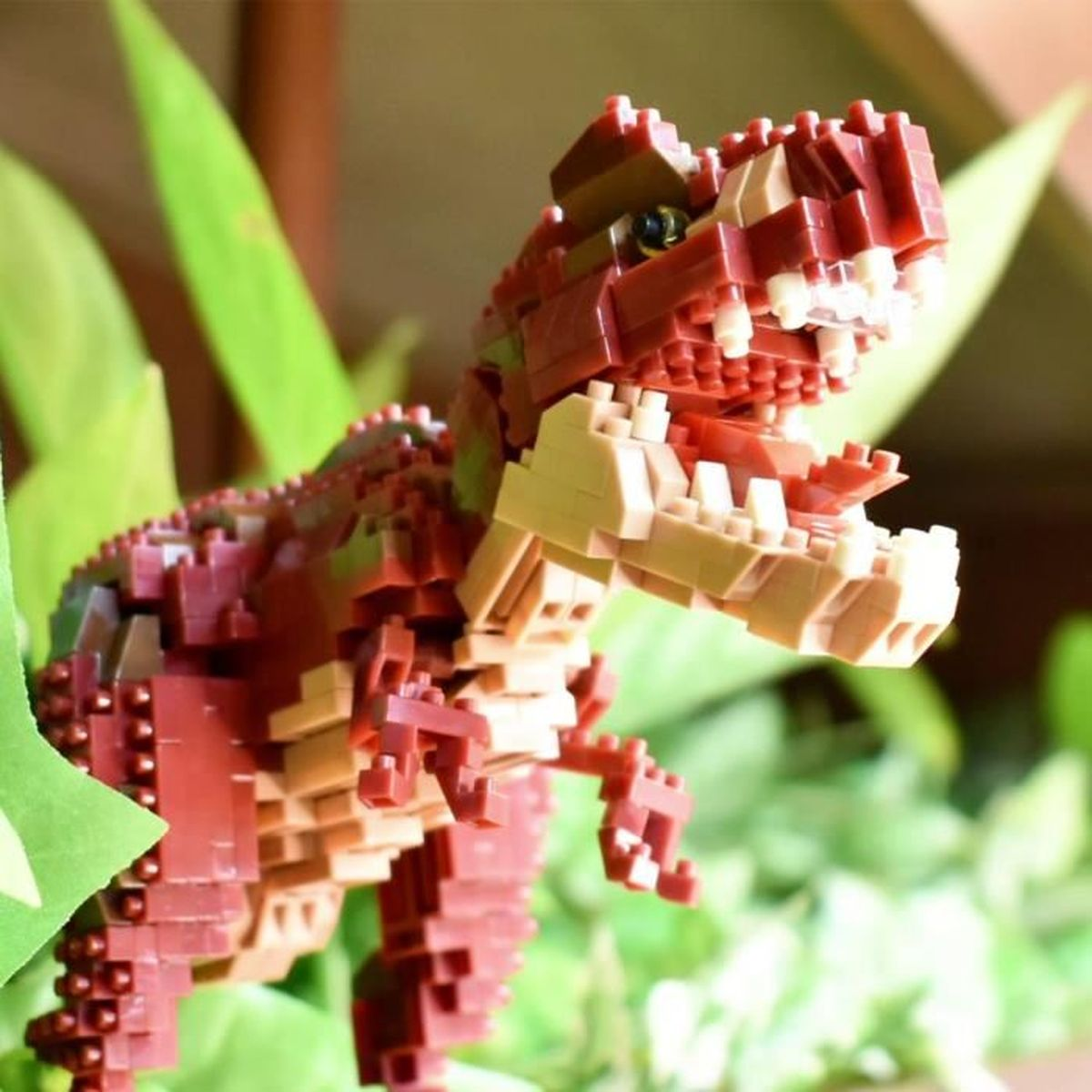 Ein aus Ziegeln gebautes Dinosaurier-Spielzeug, rot und hellbraun, mit offenem Mund. Umgeben von grünen Blättern, eine Nahaufnahme.