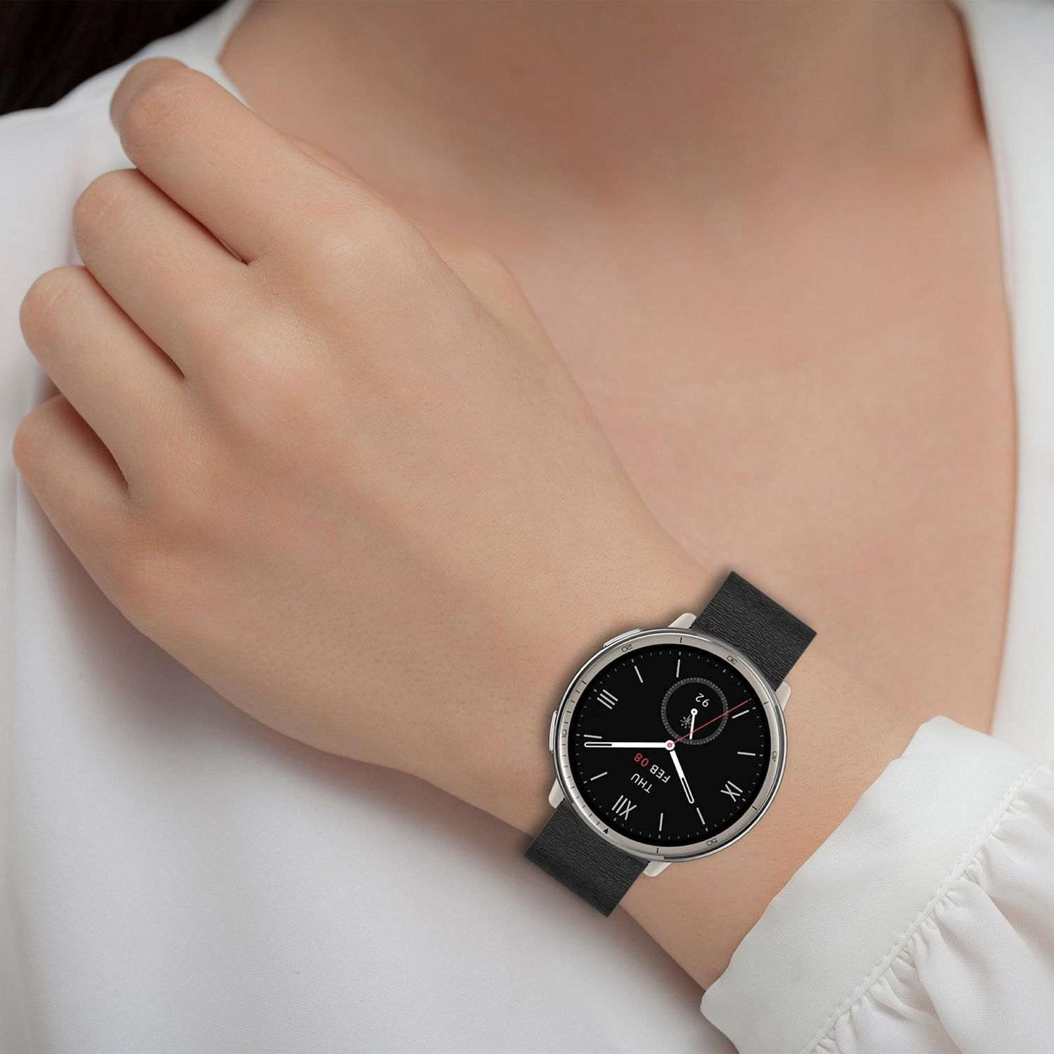 Am Handgelenk einer Frau ist eine Smartwatch mit schwarzem Armband und Zifferblatt zu sehen. Die Uhr zeigt das Datum. Die Frau trägt eine weiße Bluse.