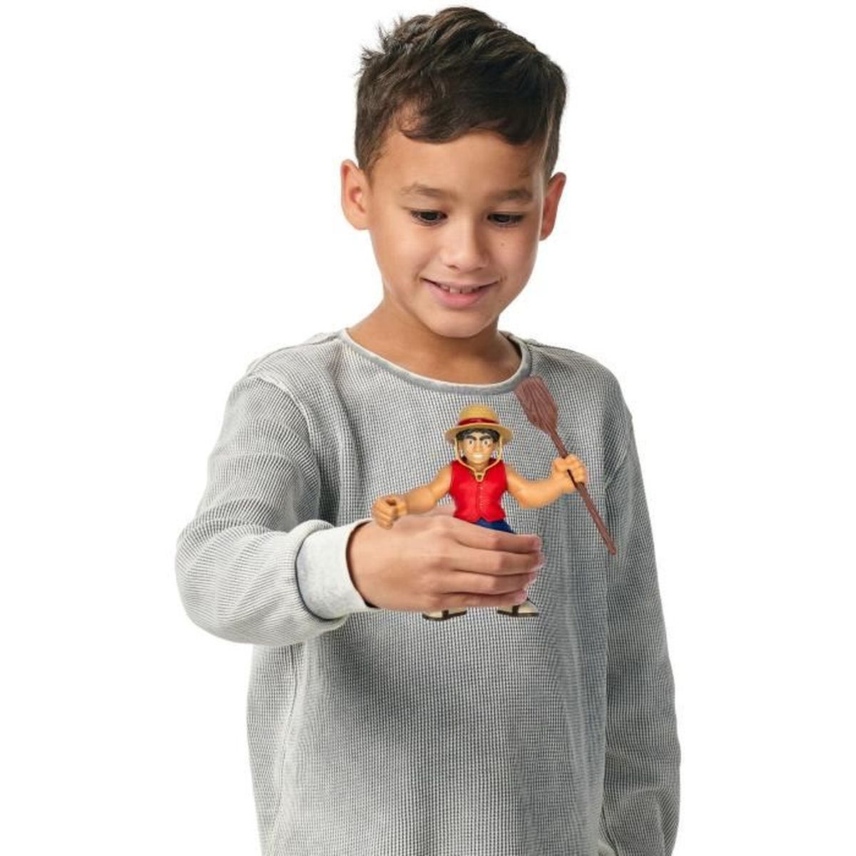 Un niño sostiene una figura de juguete de un personaje con chaleco rojo y sombrero de paja. El niño sonríe. También sostiene una paleta de madera.