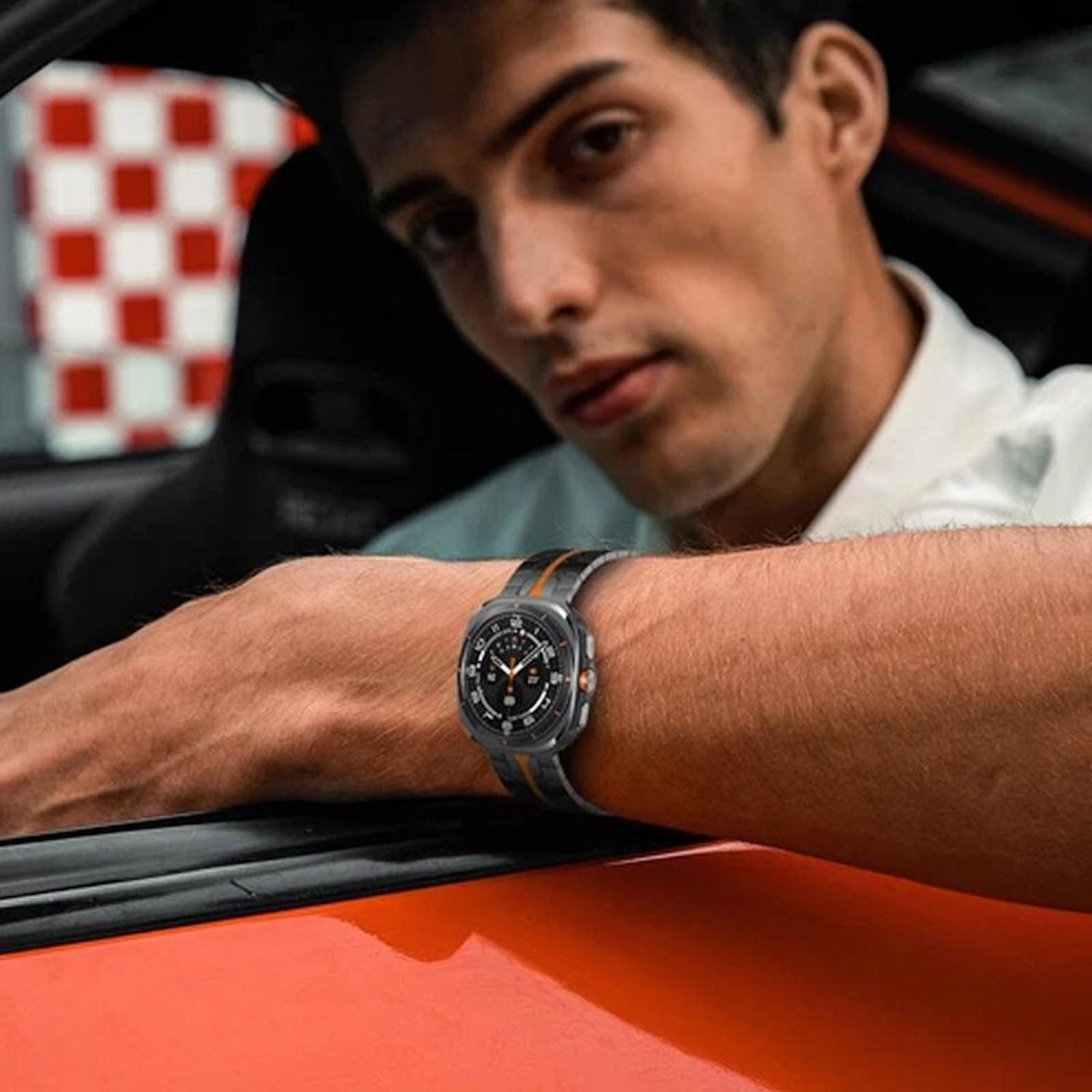 Ein Mann, der eine Uhr trägt, lehnt sich an ein Auto. Karierte Flagge sichtbar. Oranges Auto. Die Uhr hat orange und schwarze Details.