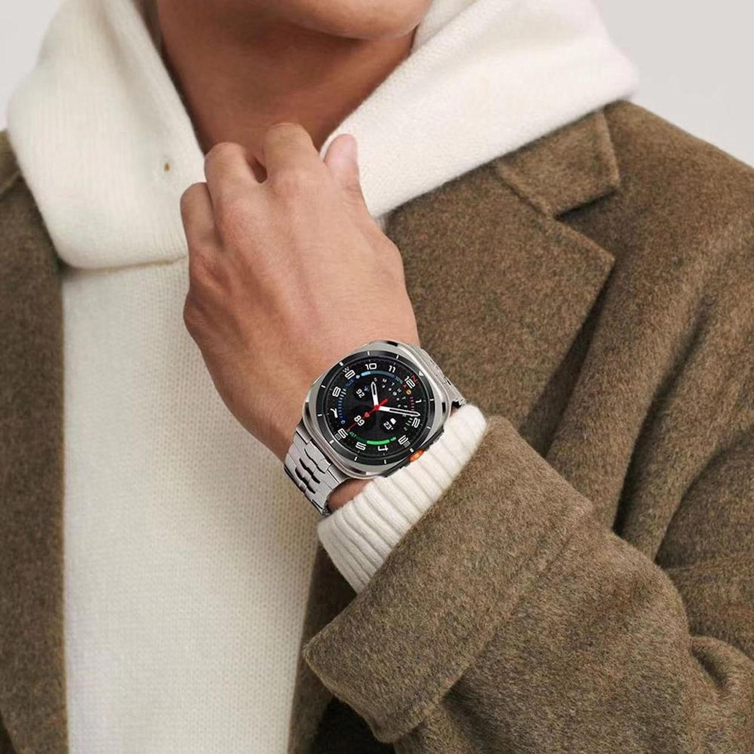 Eine Person, die eine Uhr mit silbernem Armband, einem weißen Pullover und einem braunen Mantel trägt. Das Zifferblatt ist schwarz und silber.
