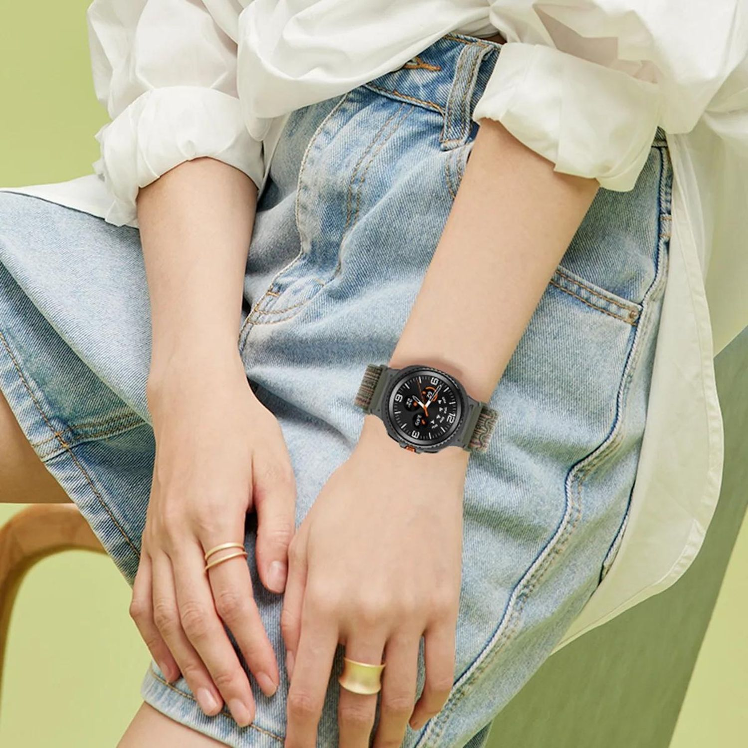 Person trägt Uhr, Ringe und Jeansshorts, weißes Hemd. Die Uhr hat ein dunkles Zifferblatt und ein grünes Band. Der Hintergrund ist grün.