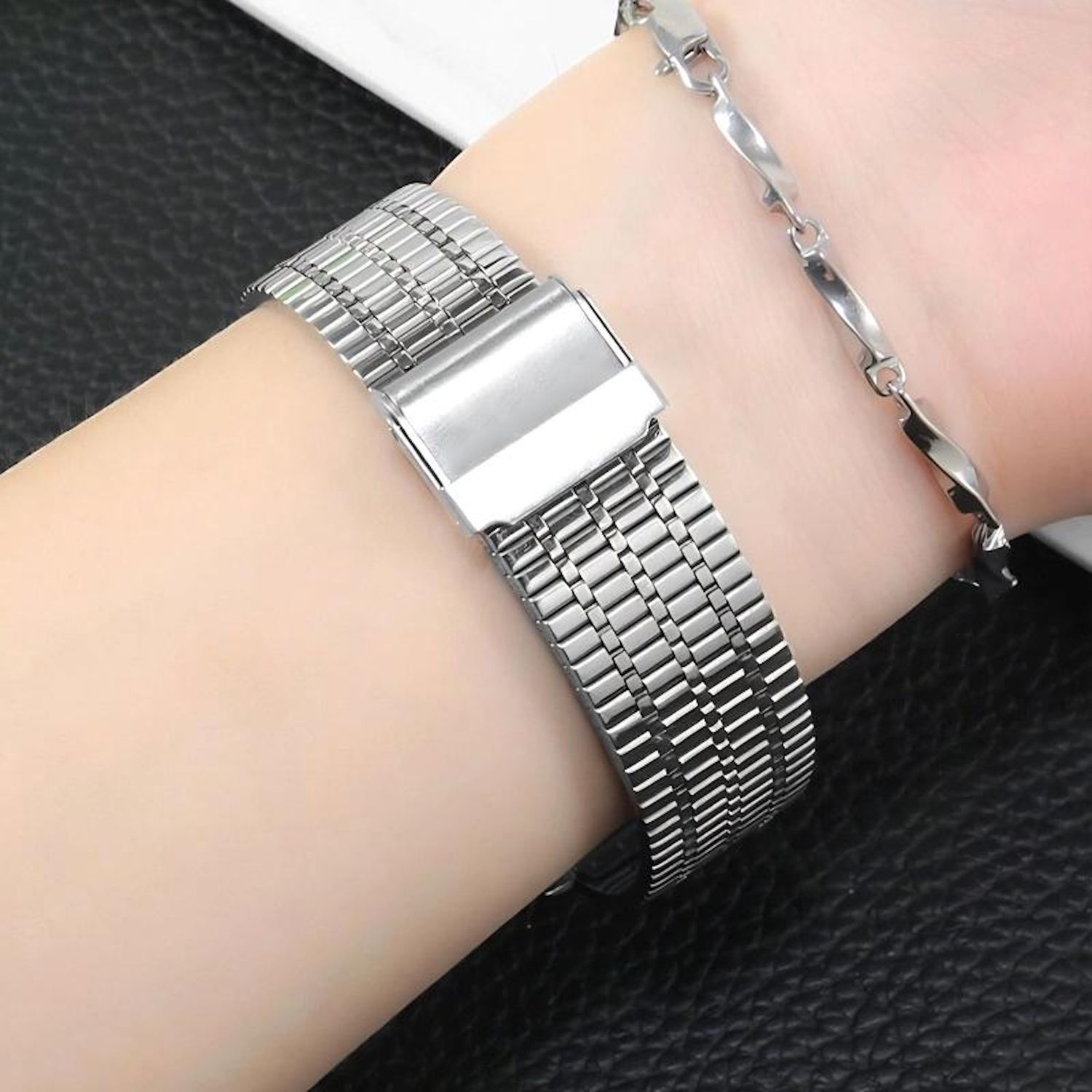 Das Handgelenk einer Person mit zwei silbernen Armbändern. Eines ist ein Uhrenarmband, das andere ein Gliederarmband.