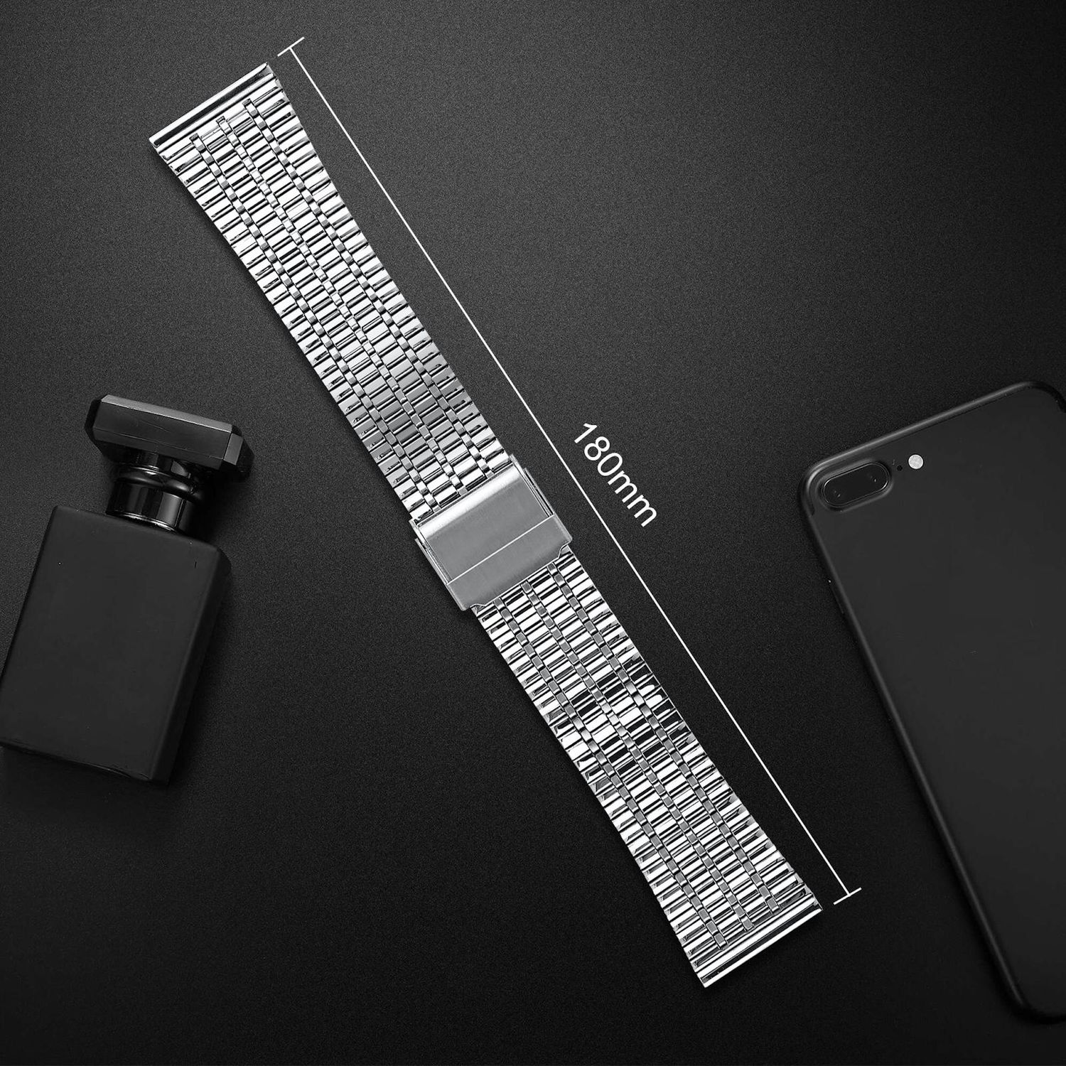 Ein silbernes Uhrenarmband, eine schwarze Parfümflasche und ein schwarzes Telefon liegen auf schwarzem Untergrund. Eine Linie zeigt 180 mm an.