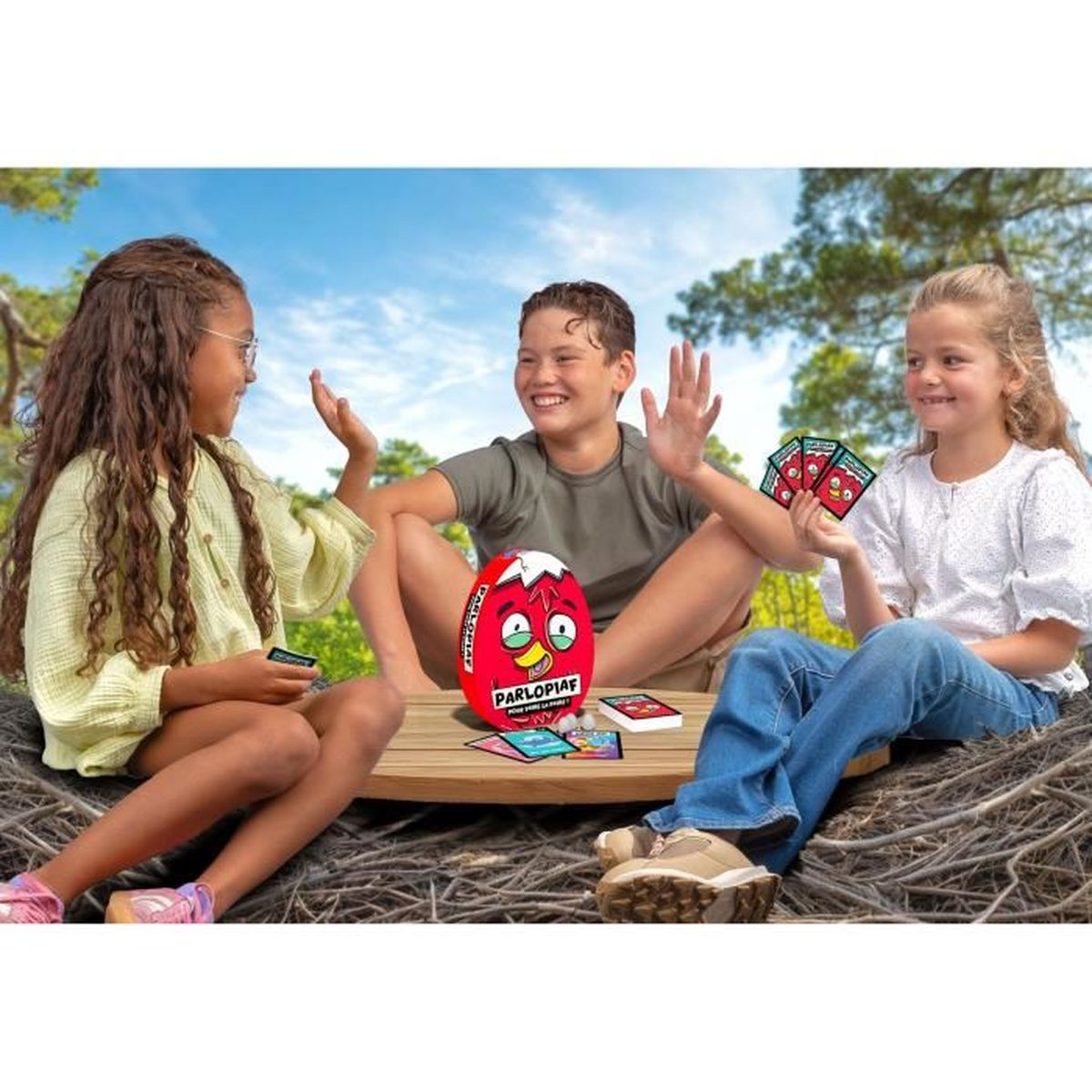 Drei Kinder spielen im Freien ein Spiel. Sie sitzen um einen Holztisch mit einem roten, eiförmigen Spiel und Karten.