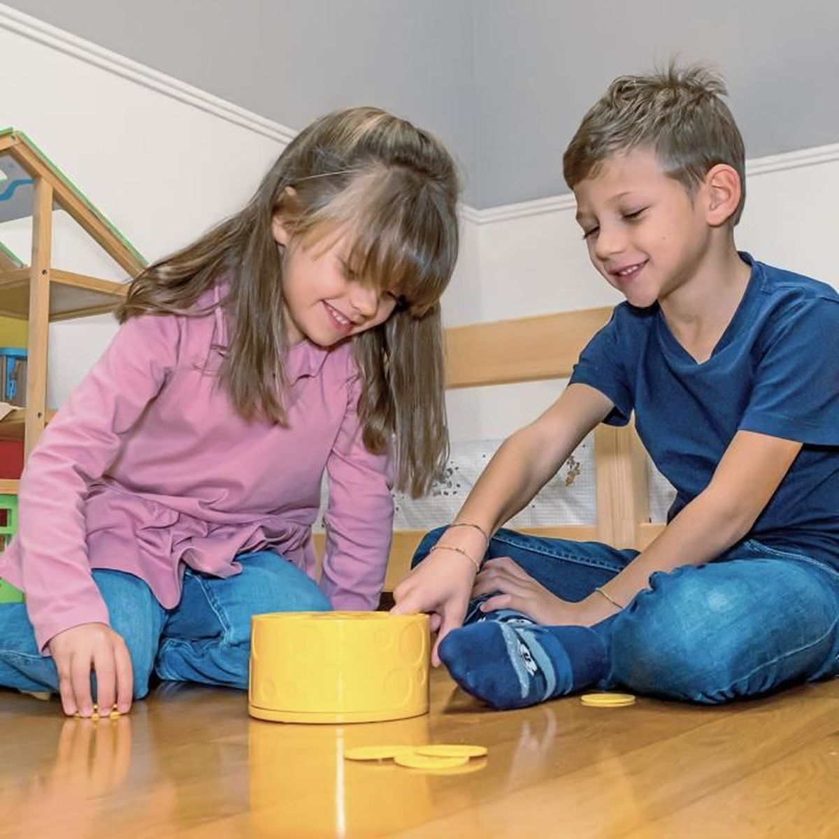 Zwei Kinder spielen ein Spiel mit gelben Scheiben und einem gelben Zylinder auf einem Holzboden. Sie lächeln und sind konzentriert.