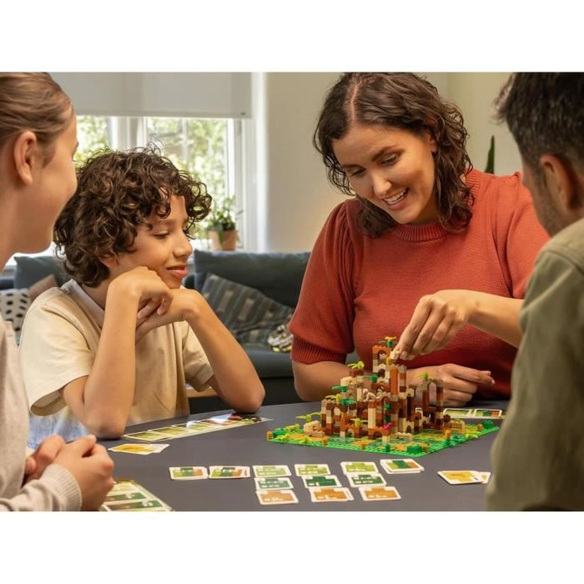 Vier mensen spelen een bordspel. Een vrouw voegt een blok toe aan een Lego-constructie. Kaarten en de constructie liggen op een grijze tafel.