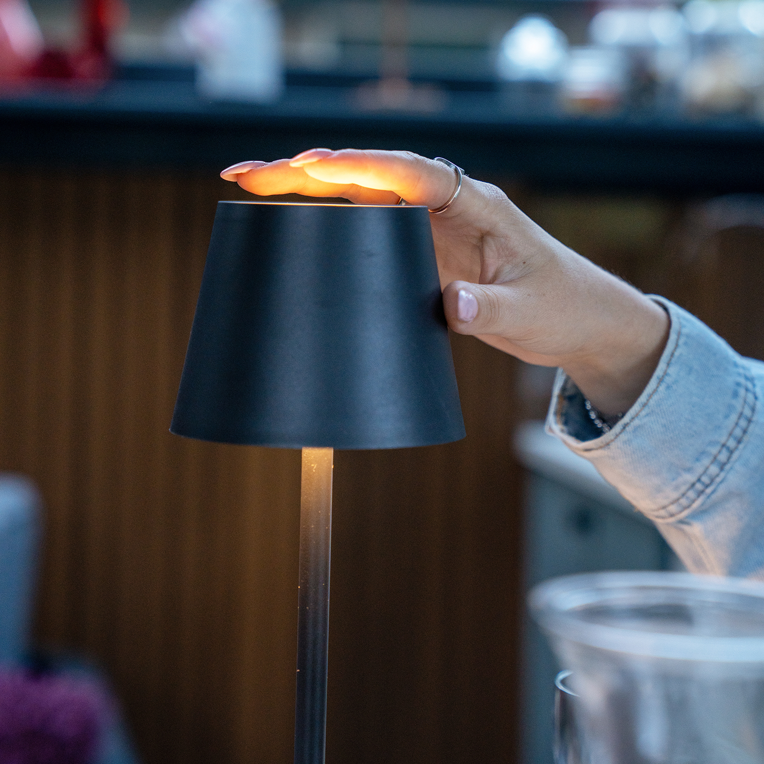 Een hand raakt een kleine zwarte lamp aan, waardoor de vingers oplichten. De achtergrond toont een vage binnenomgeving met een spijkerjasje.