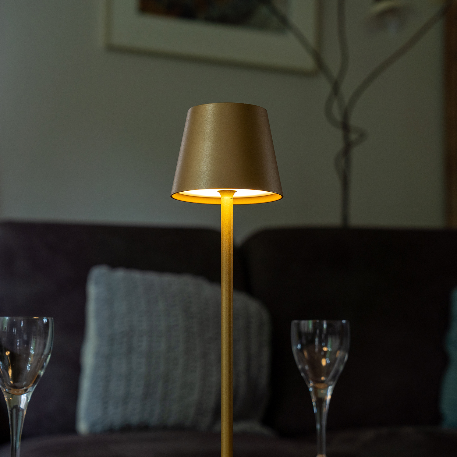Een brandende lamp met een kap, twee wijnglazen en een donkere bank op de voorgrond. Een schilderij hangt aan de muur op de achtergrond.