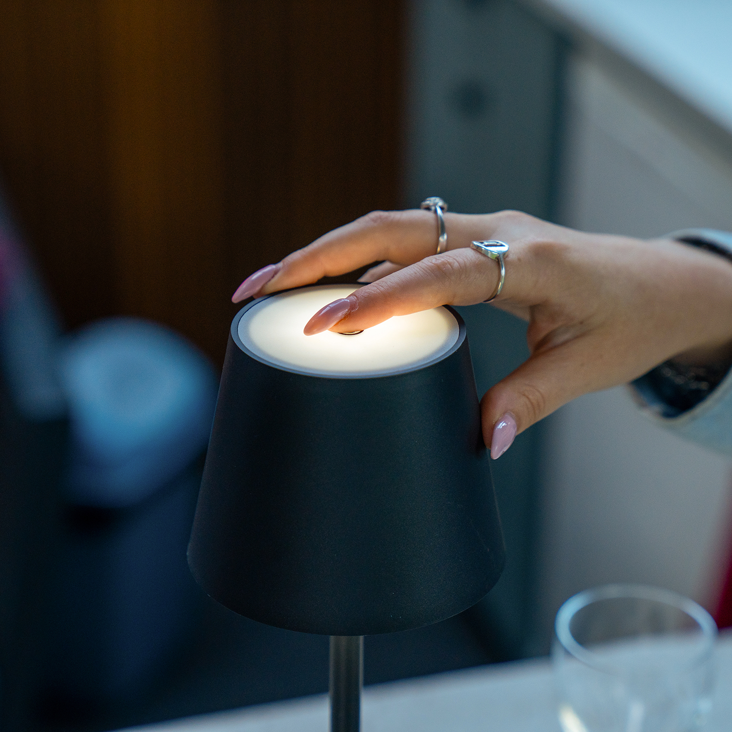 Een hand met ringen raakt de bovenkant van een zwarte lamp aan. De lamp geeft een zachte gloed, en een glas is zichtbaar op de voorgrond.