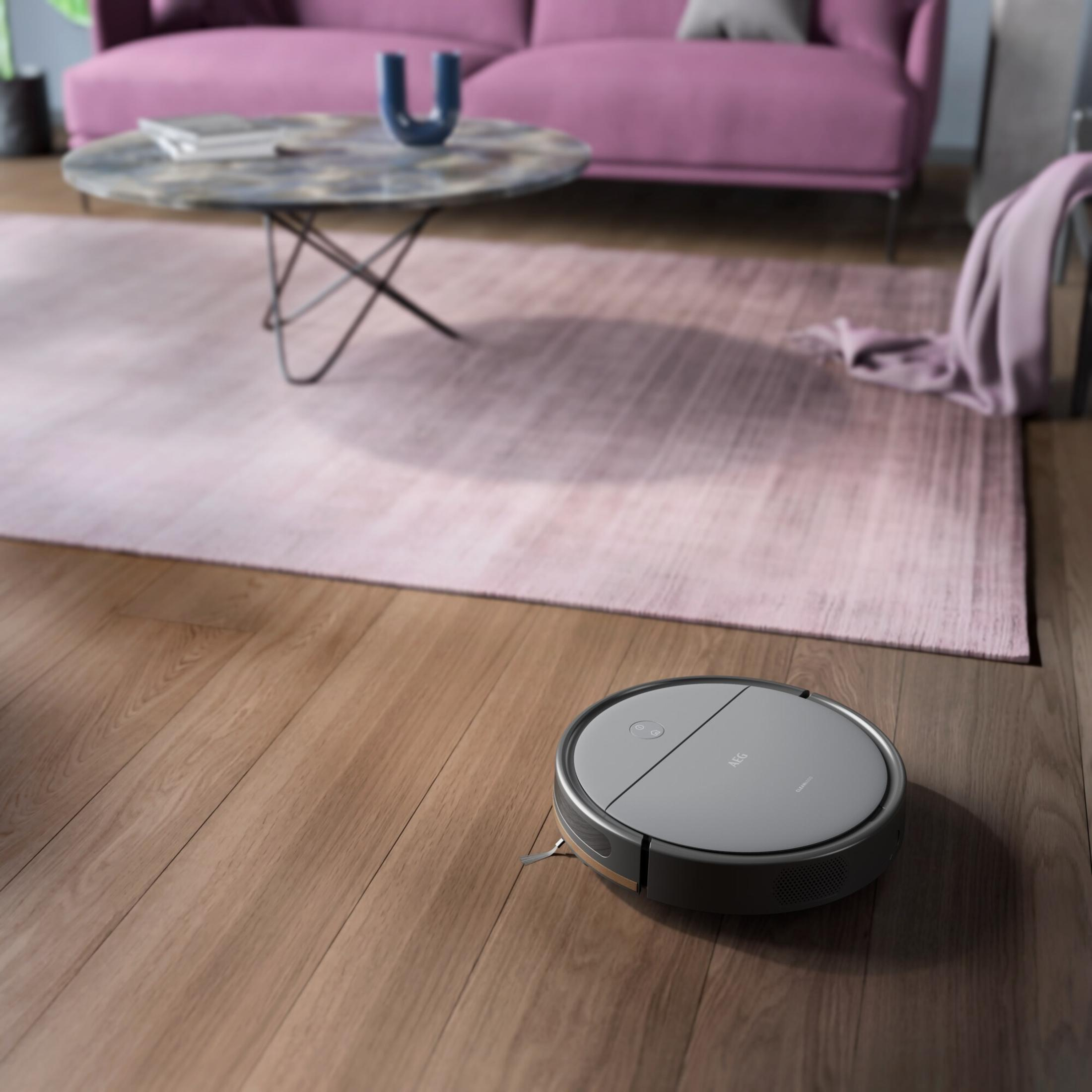 Ein Roboter-Staubsauger auf einem Holzboden. Ein rosa Teppich und Sofa befinden sich im Hintergrund, zusammen mit einem Couchtisch.