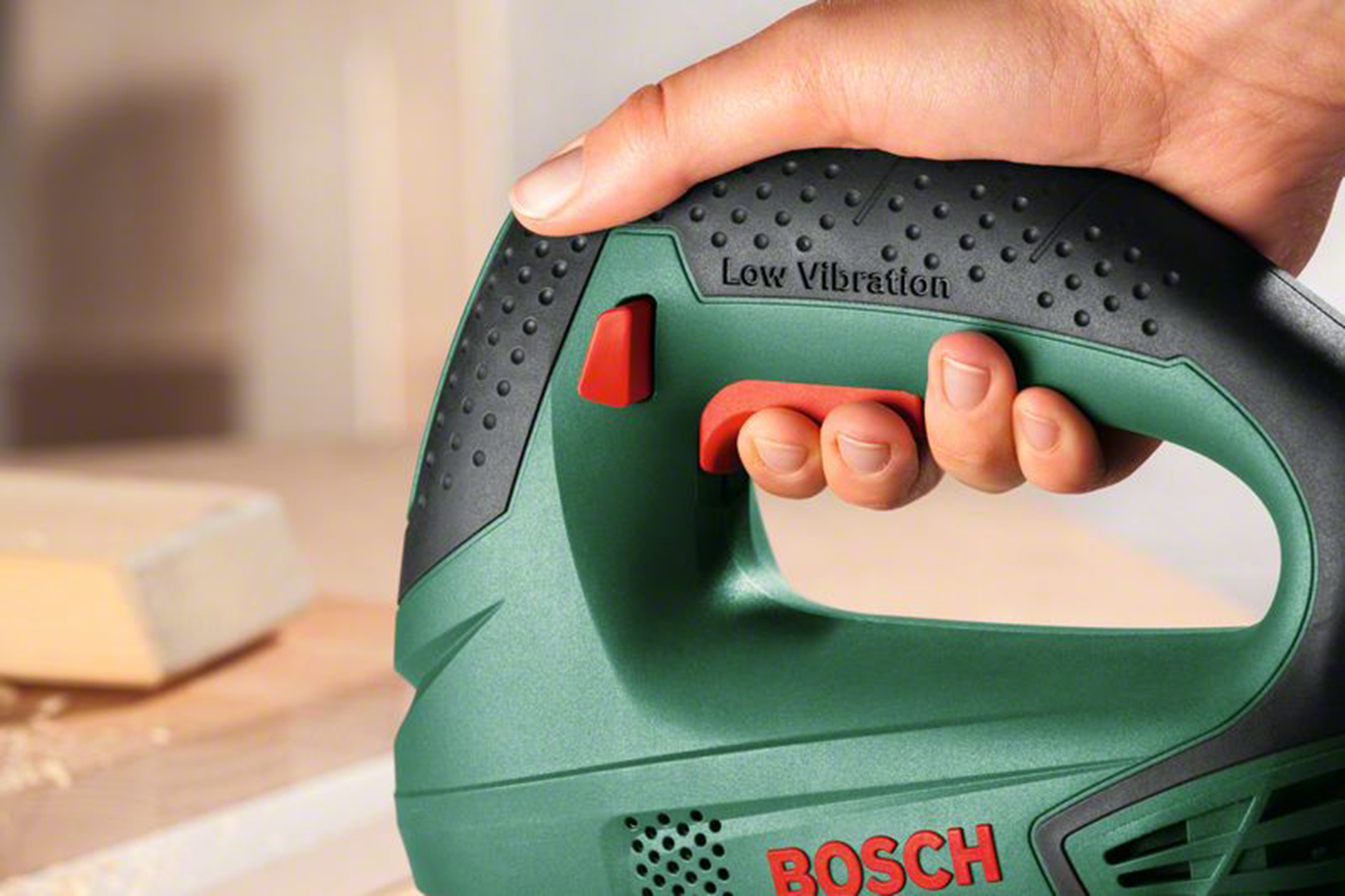 Hand die een groene en zwarte Bosch decoupeerzaag vasthoudt met rode accenten. 'Low Vibration' is zichtbaar. Een houten blok staat op de achtergrond.