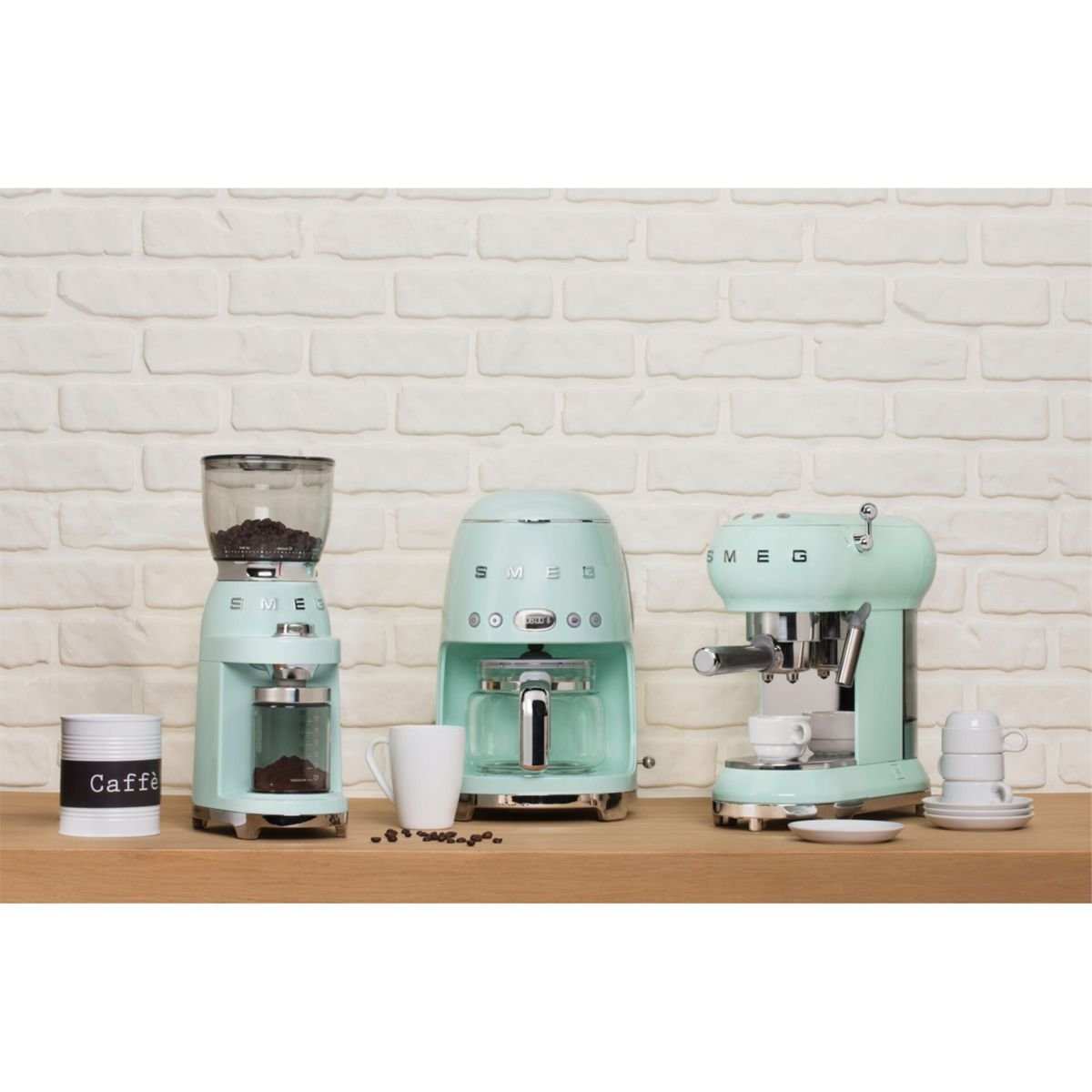 Máquinas y accesorios de café color menta sobre una estantería de madera contra una pared de ladrillos blancos. Incluye un molinillo, una cafetera y una máquina de espresso.