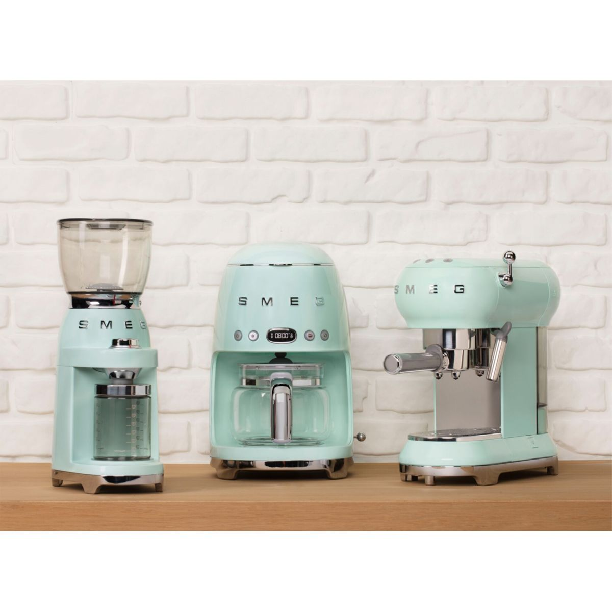 Tres cafeteras SMEG verde menta sobre una superficie de madera contra una pared de ladrillos blancos. De izquierda: molinillo, cafetera, máquina de espresso.