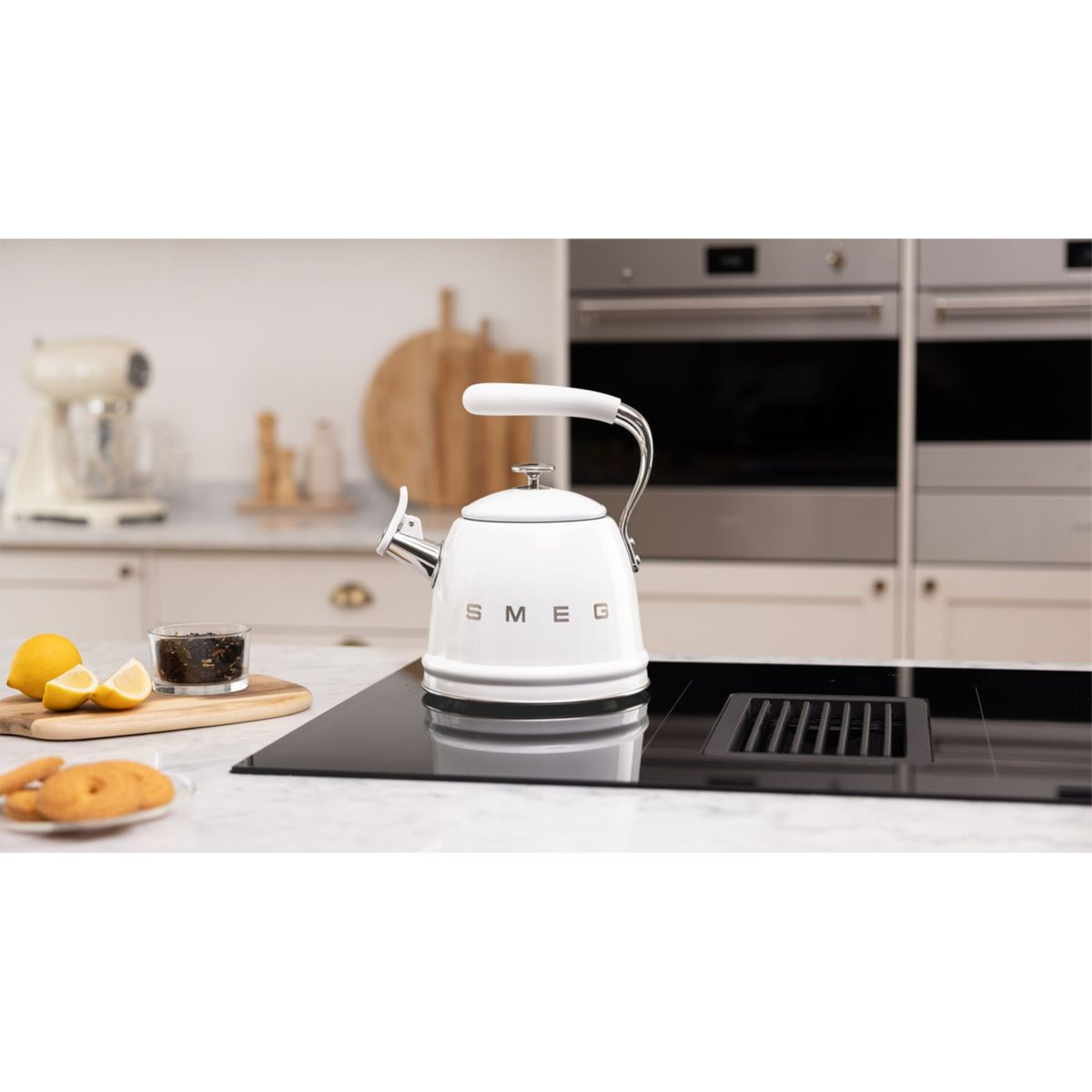 Una tetera Smeg blanca está sobre una estufa negra. Hay limones, té y galletas cerca. Electrodomésticos de cocina en el fondo.