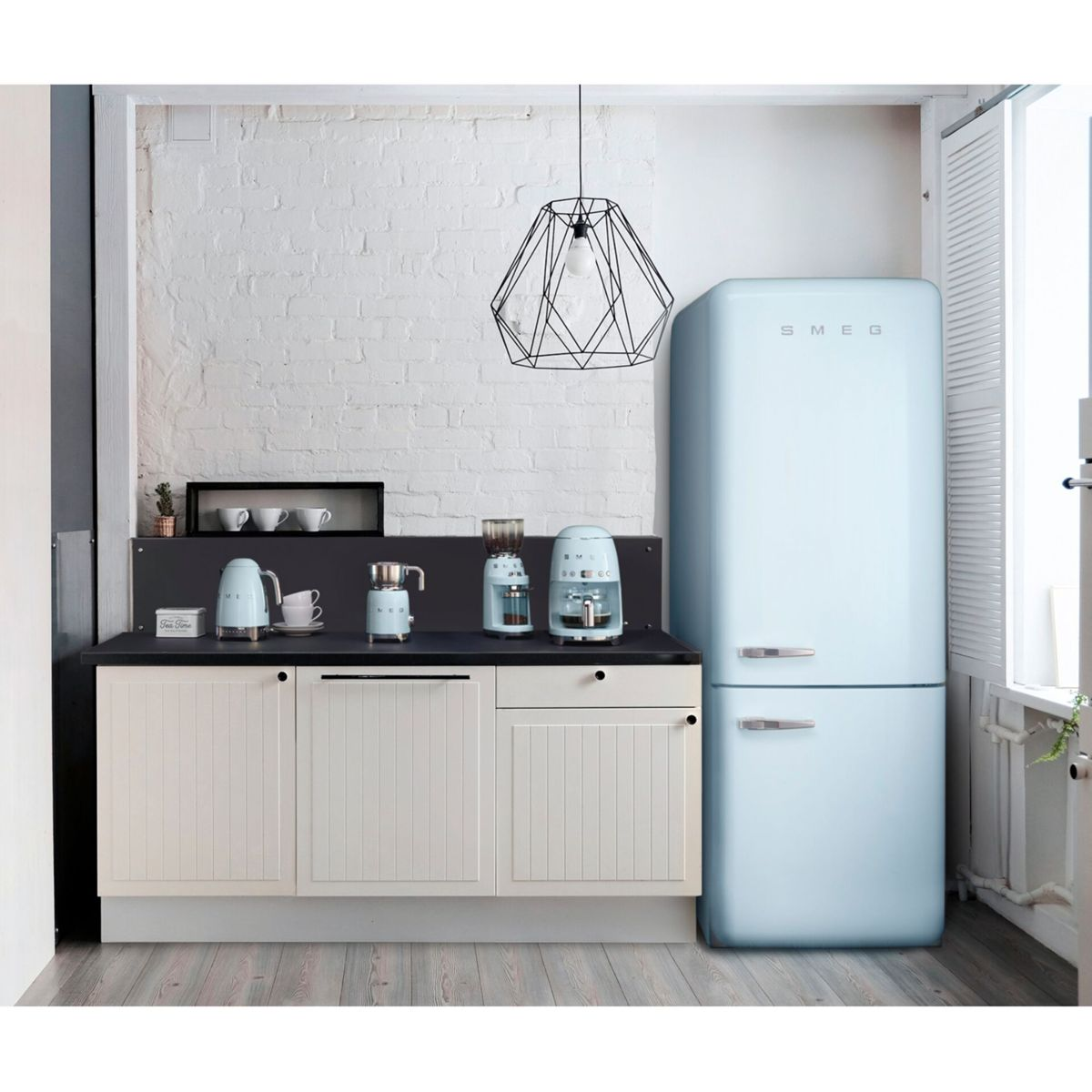 Une cuisine avec un réfrigérateur Smeg bleu, des machines à café et des placards blancs. Une lampe géométrique noire est suspendue.