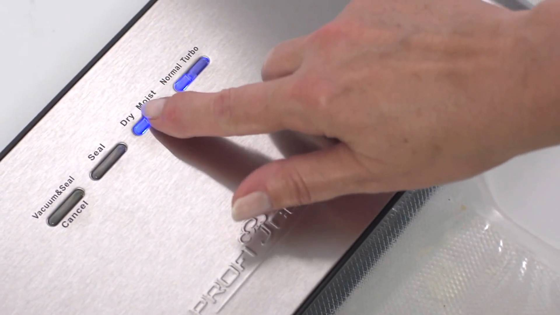 Ręka naciska przycisk 'Dry' na zgrzewarce próżniowej. Maszyna jest srebrna, z podświetlanymi opcjami. Widoczna jest plastikowa torba.