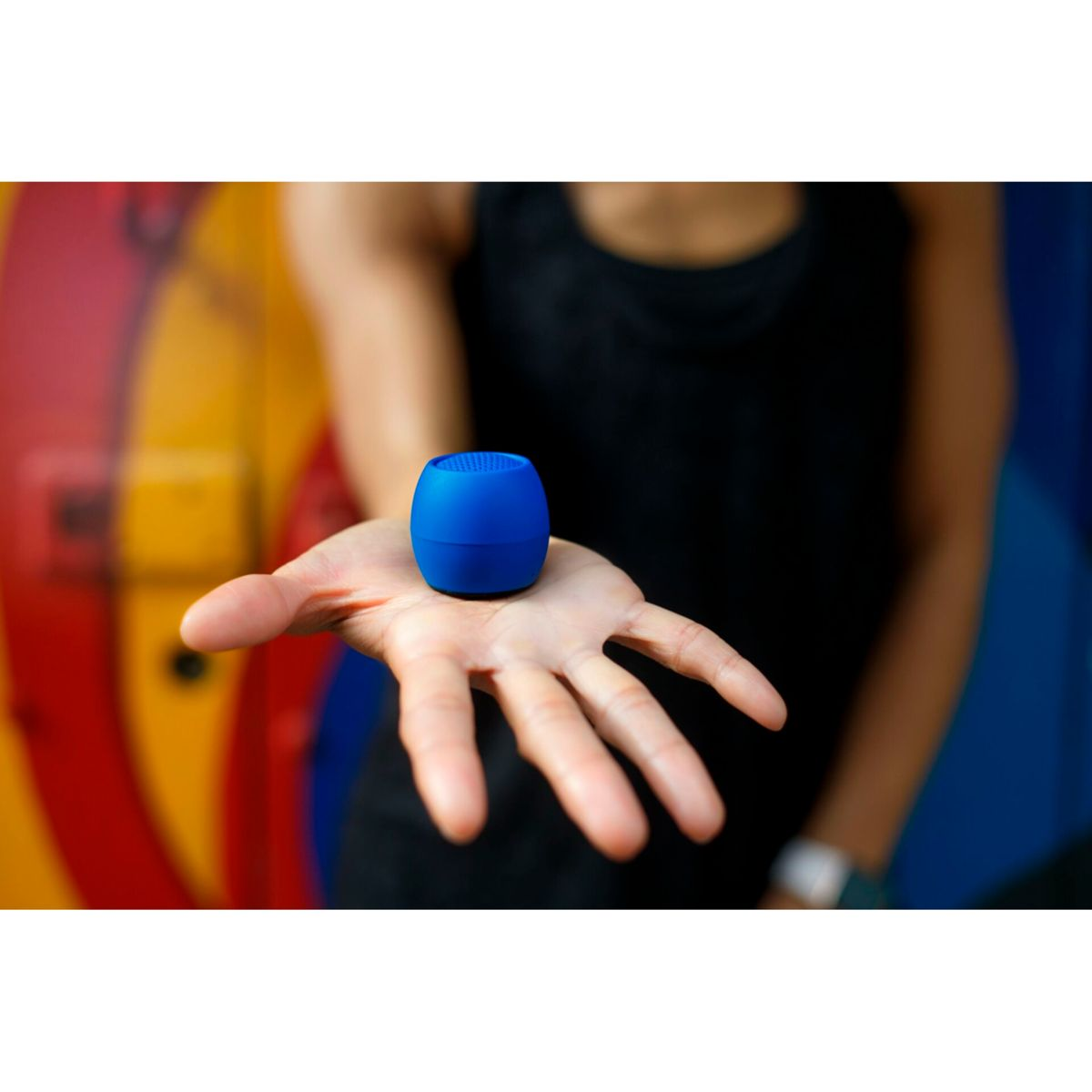 Otwarta dłoń trzyma mały, niebieski, cylindryczny głośnik. Tło to kolorowy wzór przypominający tarczę.
