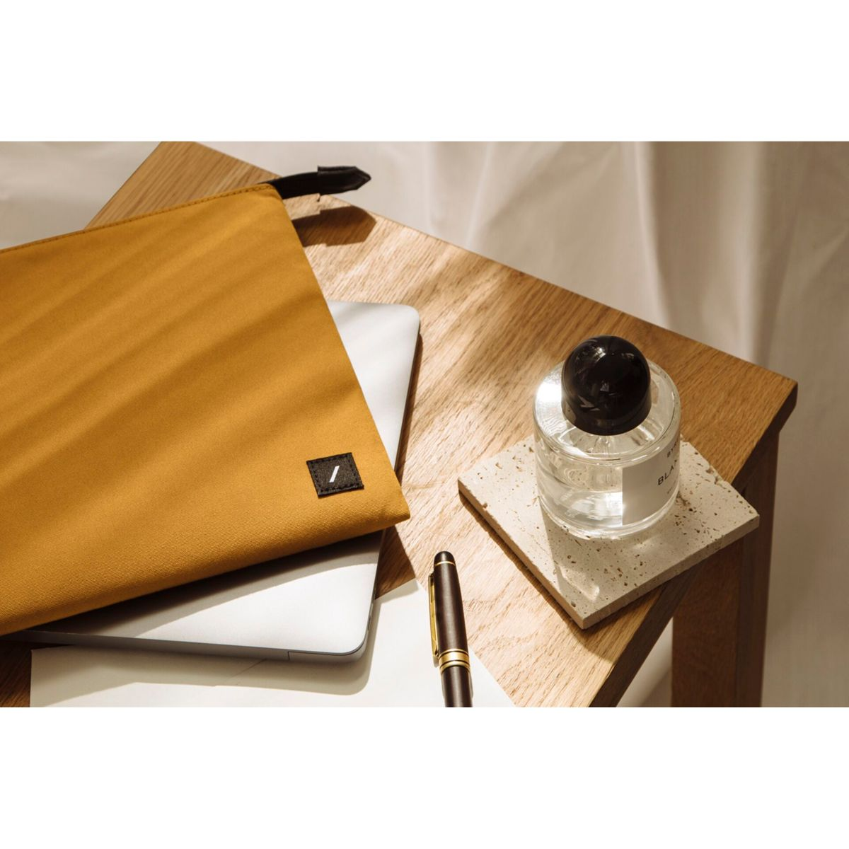Une table en bois avec un ordinateur portable, une pochette moutarde, un flacon de parfum, un stylo et un dessous de verre. Lumière douce et fond blanc.