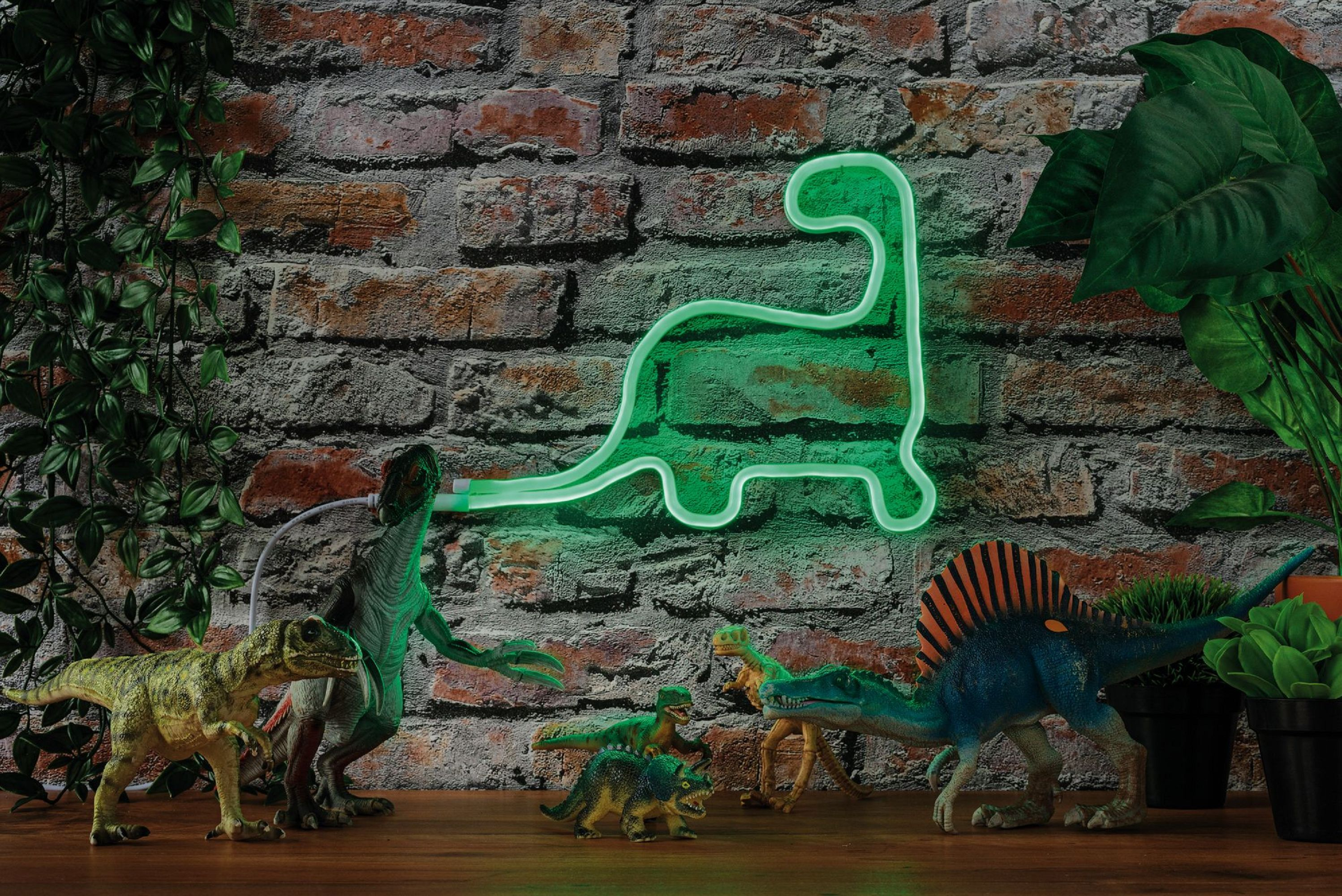 Ein grünes Neon-Dinosaurierschild an einer Backsteinmauer mit Spielzeugdinosauriern auf einer Holzoberfläche und Pflanzen.