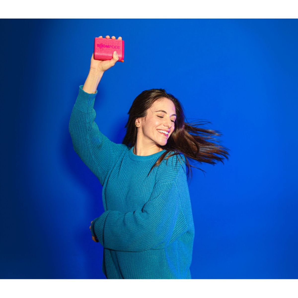 Frau in blauem Pullover hält rosa Objekt, lächelt. Blauer Hintergrund. Lange braune Haare. Das Objekt hat das Wort BOOMPODS.