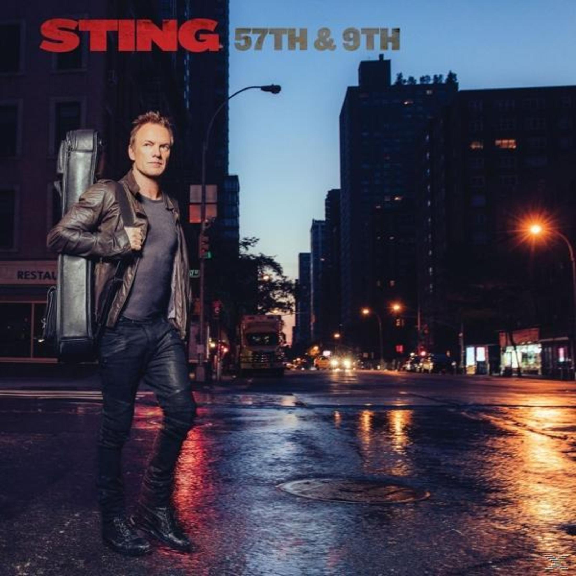Sting houdt een gitaarkoffer vast, staand op een stadsstraat 's nachts. Gebouwen en straatverlichting op de achtergrond.