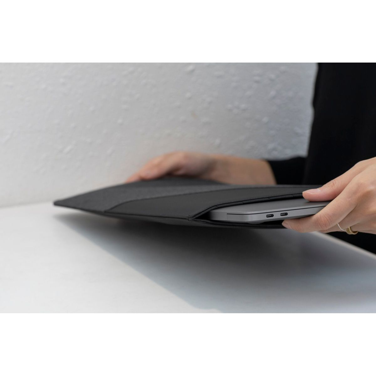 Una persona tiene in mano una custodia nera per laptop con un laptop all'interno. La custodia è appoggiata su una superficie bianca, contro un muro strutturato.