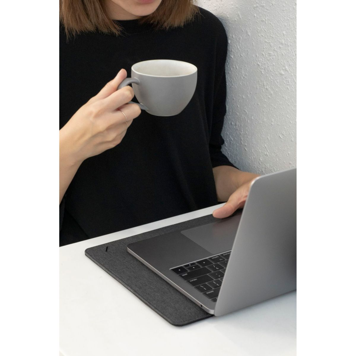 Una persona in nero tiene una tazza grigia e usa un laptop su una scrivania bianca. Il laptop è appoggiato su un tappetino grigio.