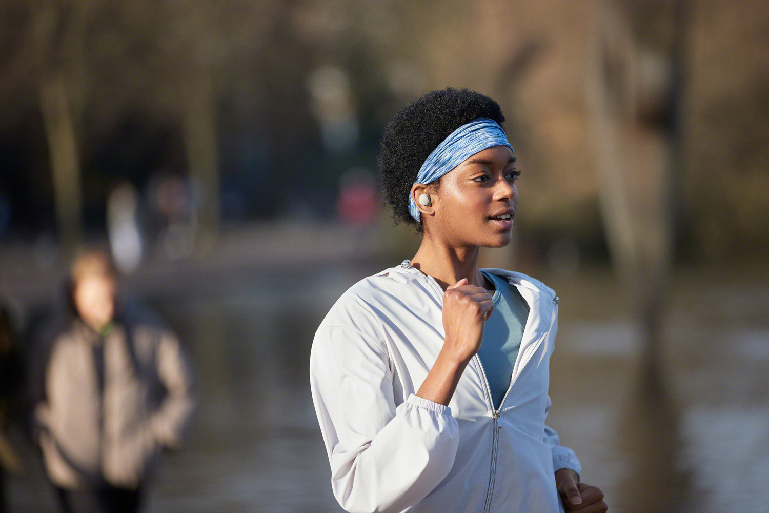 Eine Frau joggt im Freien, trägt ein blaues Stirnband und Ohrhörer. Sie trägt eine weiße Jacke, im Hintergrund ist ein Park.