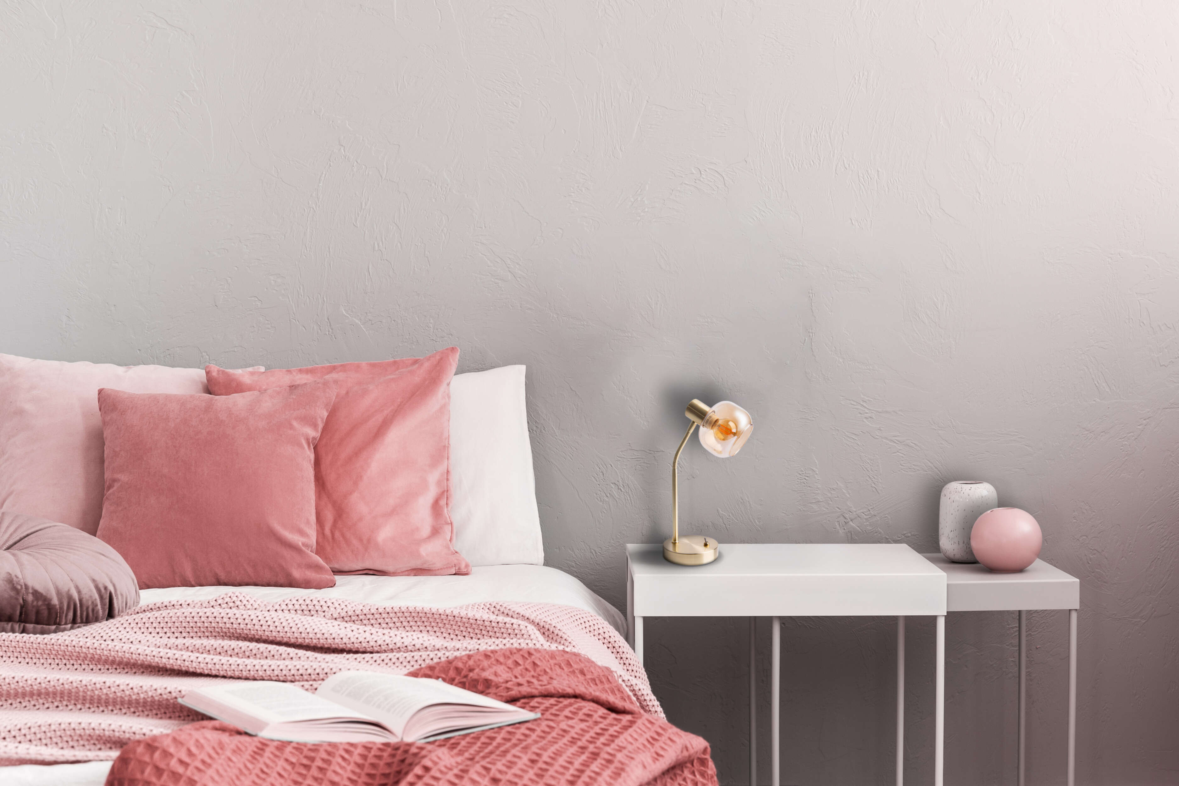 Schlafzimmer mit rosa Bettwäsche, Kissen und einem Buch. Eine Lampe und Vasen stehen auf einem weißen Beistelltisch.