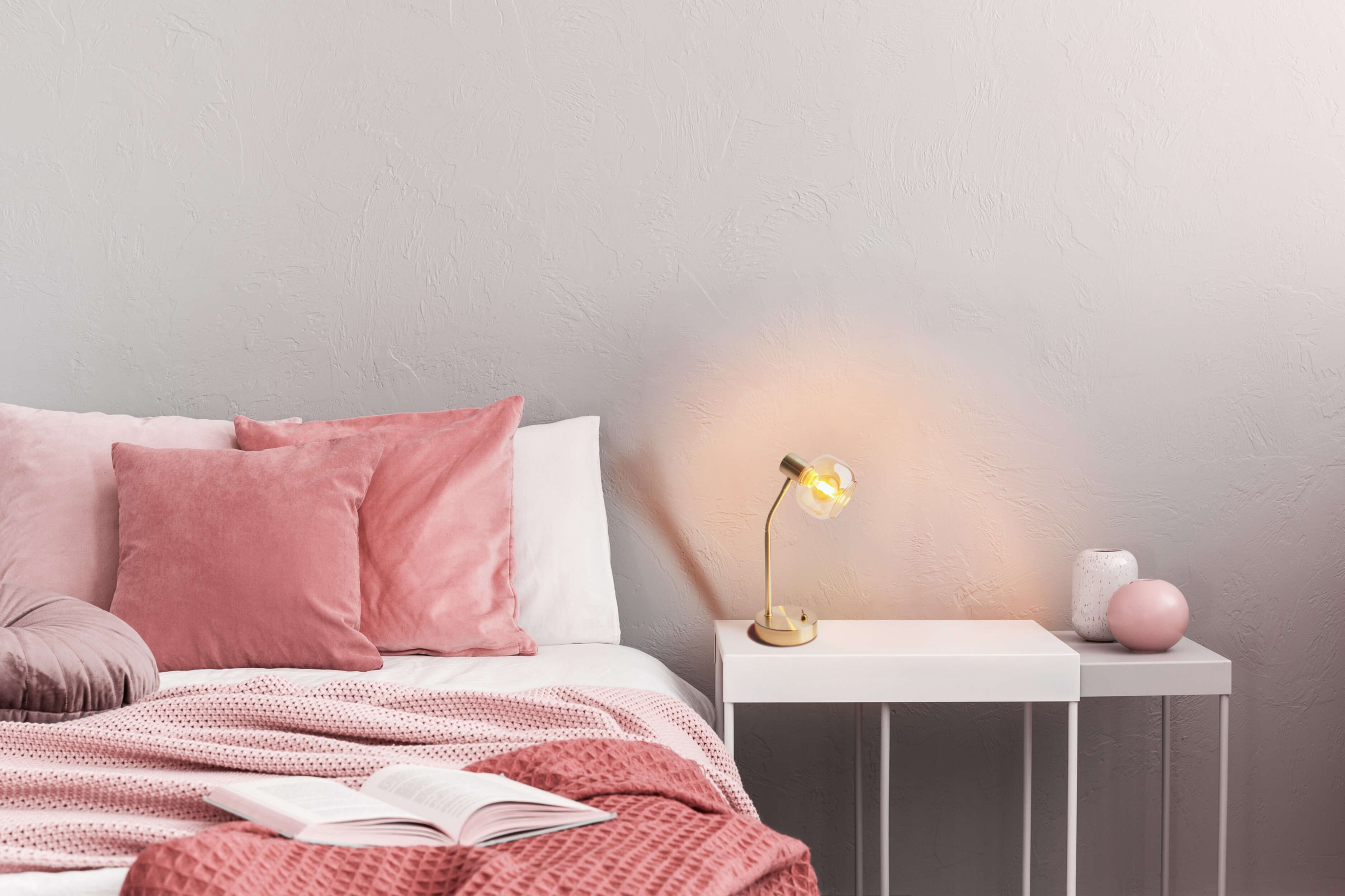 Schlafzimmer mit Bett, Kissen, Decke, Buch, Lampe und Vasen. Farben: Rosa, Weiß und Gold. Hintergrund ist graue Wand.
