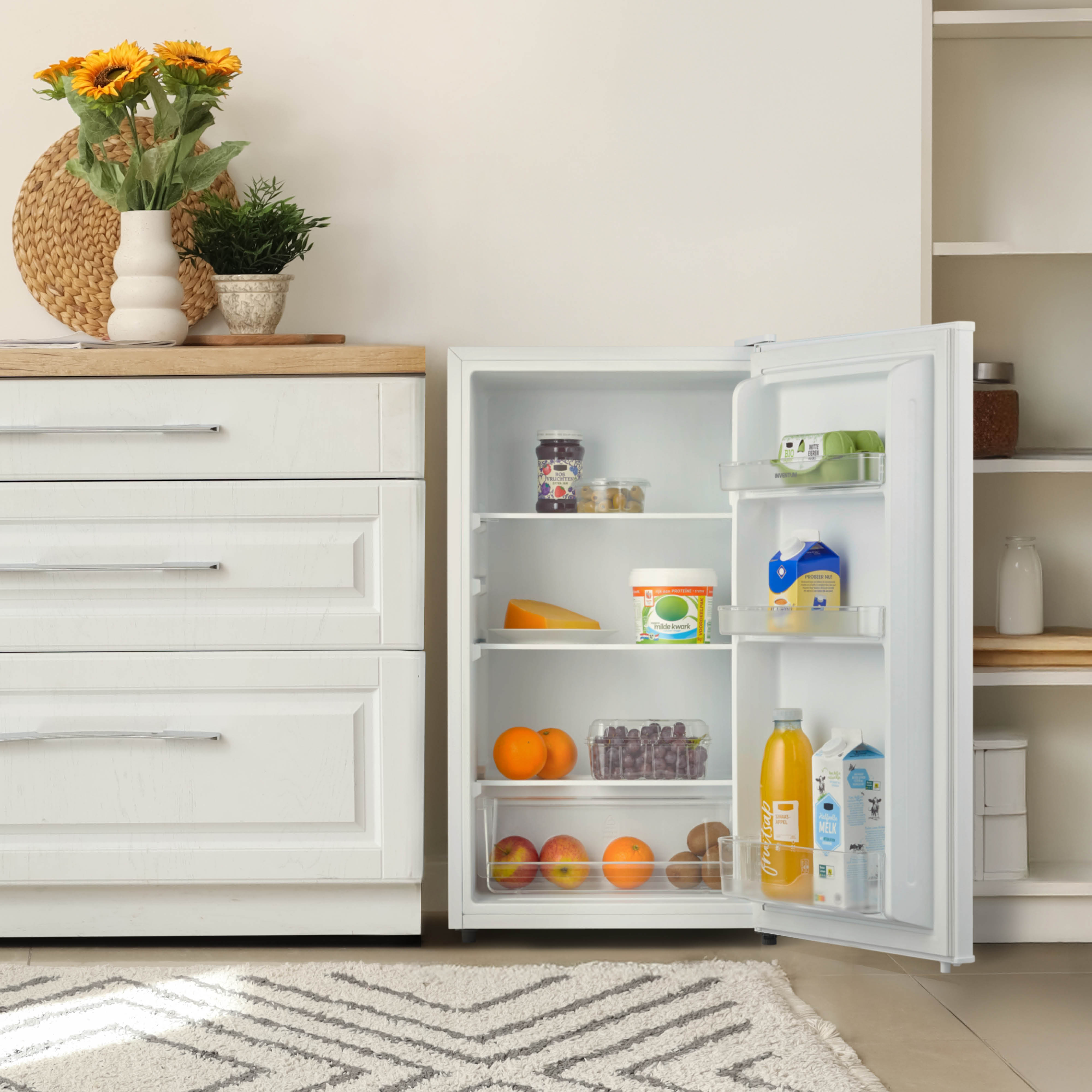 A refrigerator with open door, filled with fruits, cheese, and drinks. Sunflowers in a vase are on the counter.