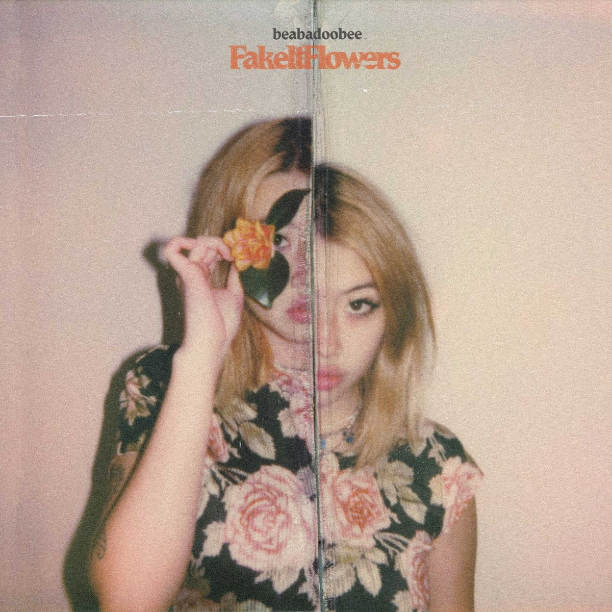 Donna a due facce che tiene un fiore. Indossa un top floreale con sfondo chiaro. Il nome dell'album "Fake It Flowers" è in alto.