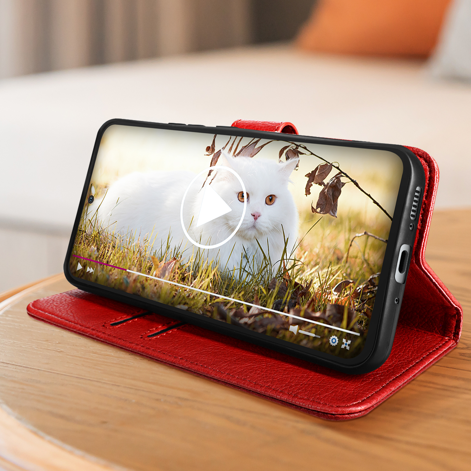 Ein Telefon zeigt ein Video einer weißen Katze im Gras. Das Telefon befindet sich in einer roten Lederhülle auf einer Holzoberfläche.
