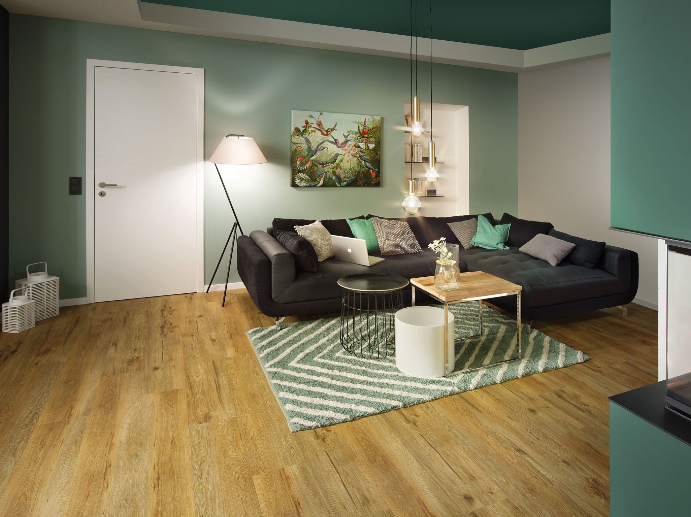 Ein modernes Wohnzimmer mit schwarzem Sofa, Holzboden und grünen Wänden. Es gibt einen Teppich, eine Lampe und Kunstwerke.