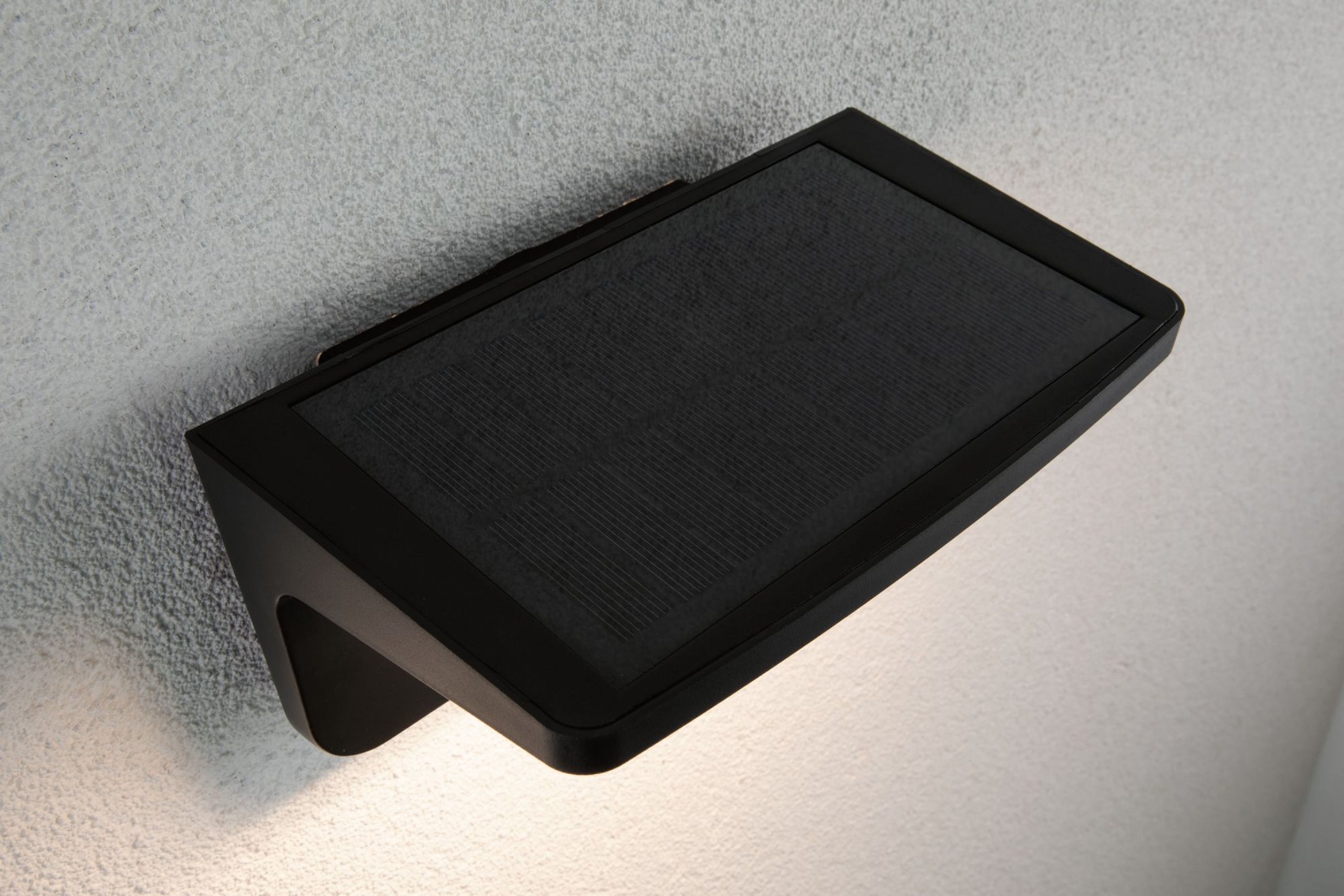 Czarna lampa ścienna zasilana energią słoneczną jest zamontowana na teksturowanej, białawej ścianie. Światło jest włączone.